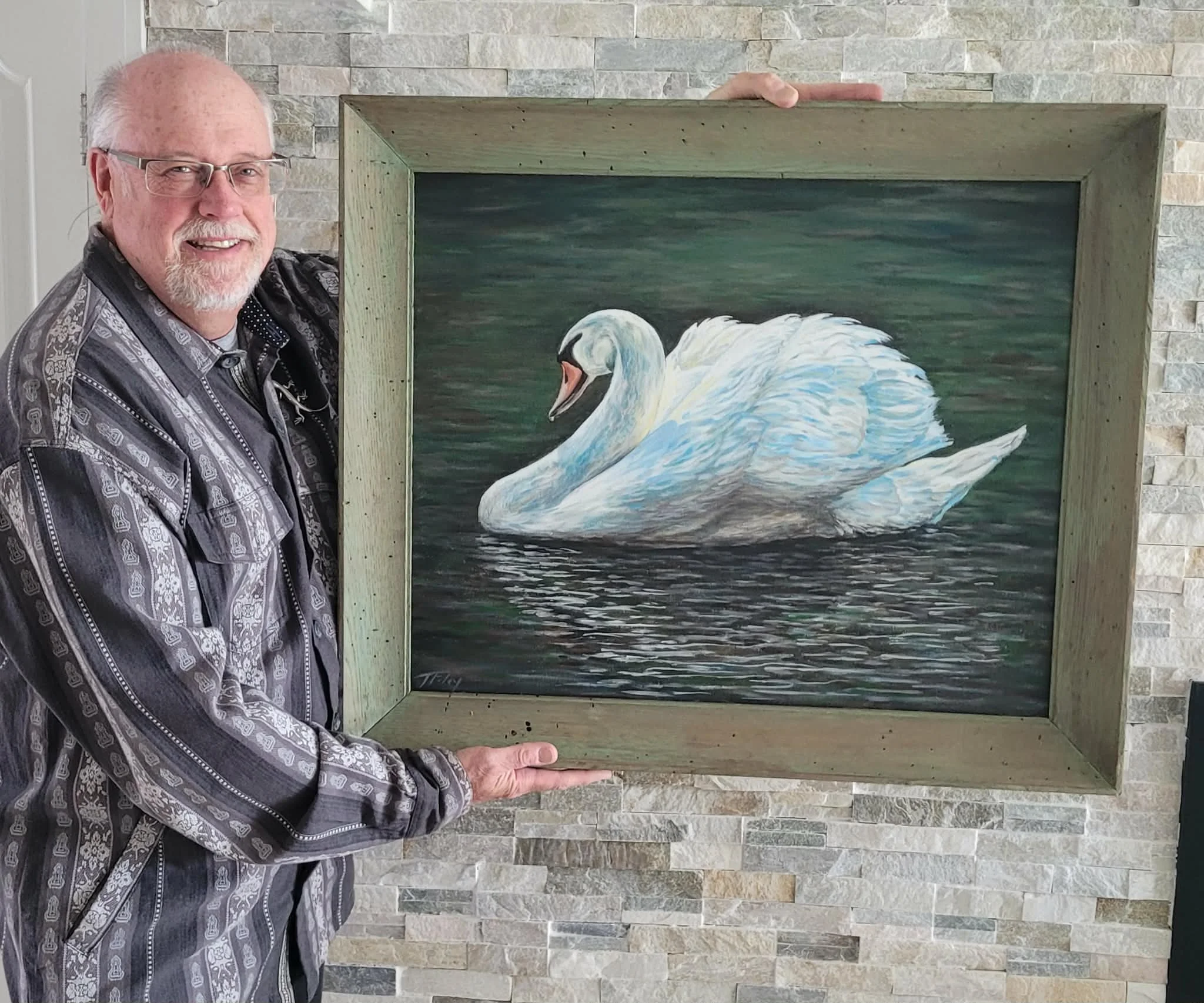 An older man with glasses and a gray beard is holding a framed painting of a white swan on water, standing in front of a stone wall.