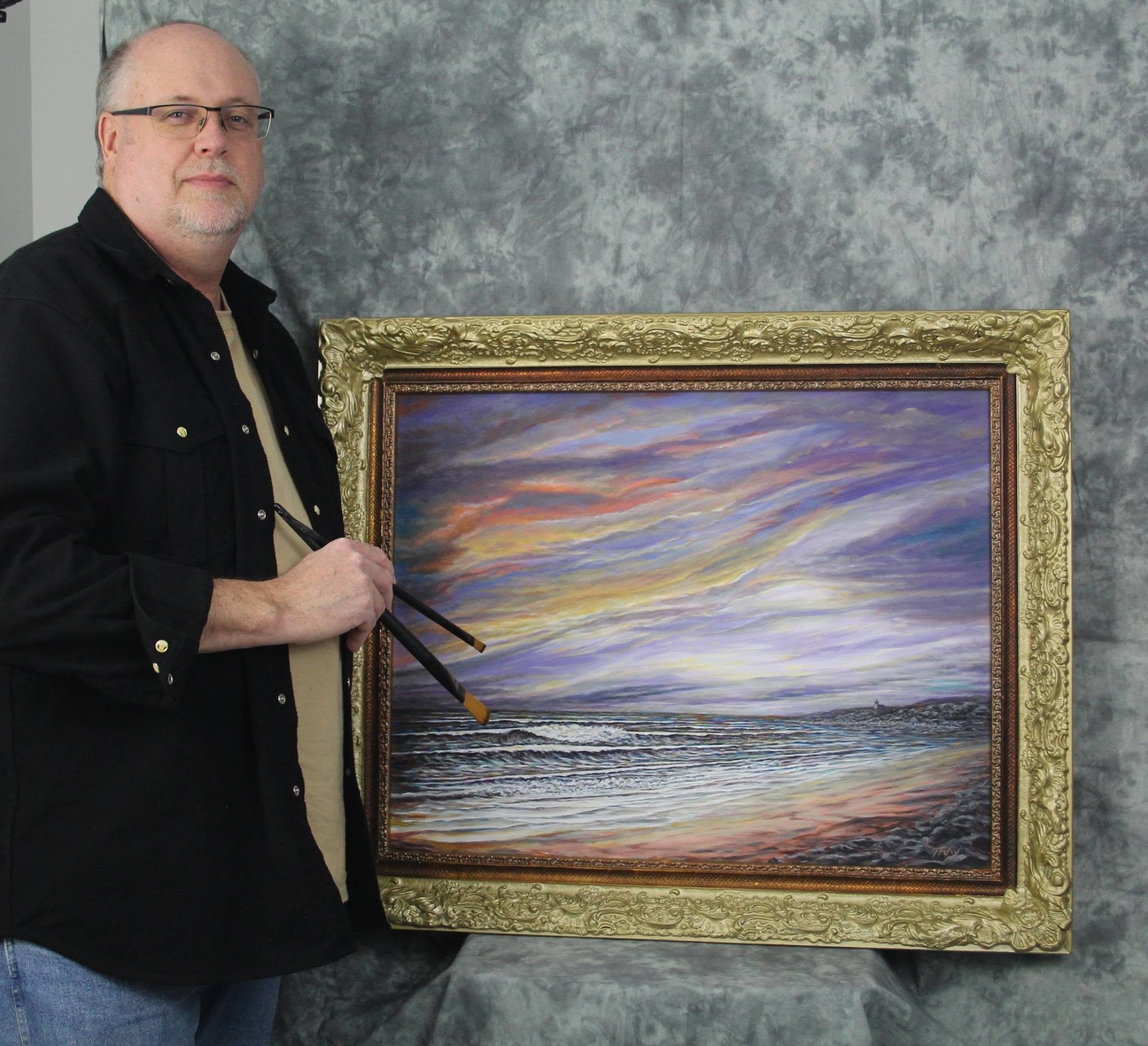 A man with glasses and a beard, wearing a black shirt, sitting beside a framed painting of a colorful sunset over the ocean.