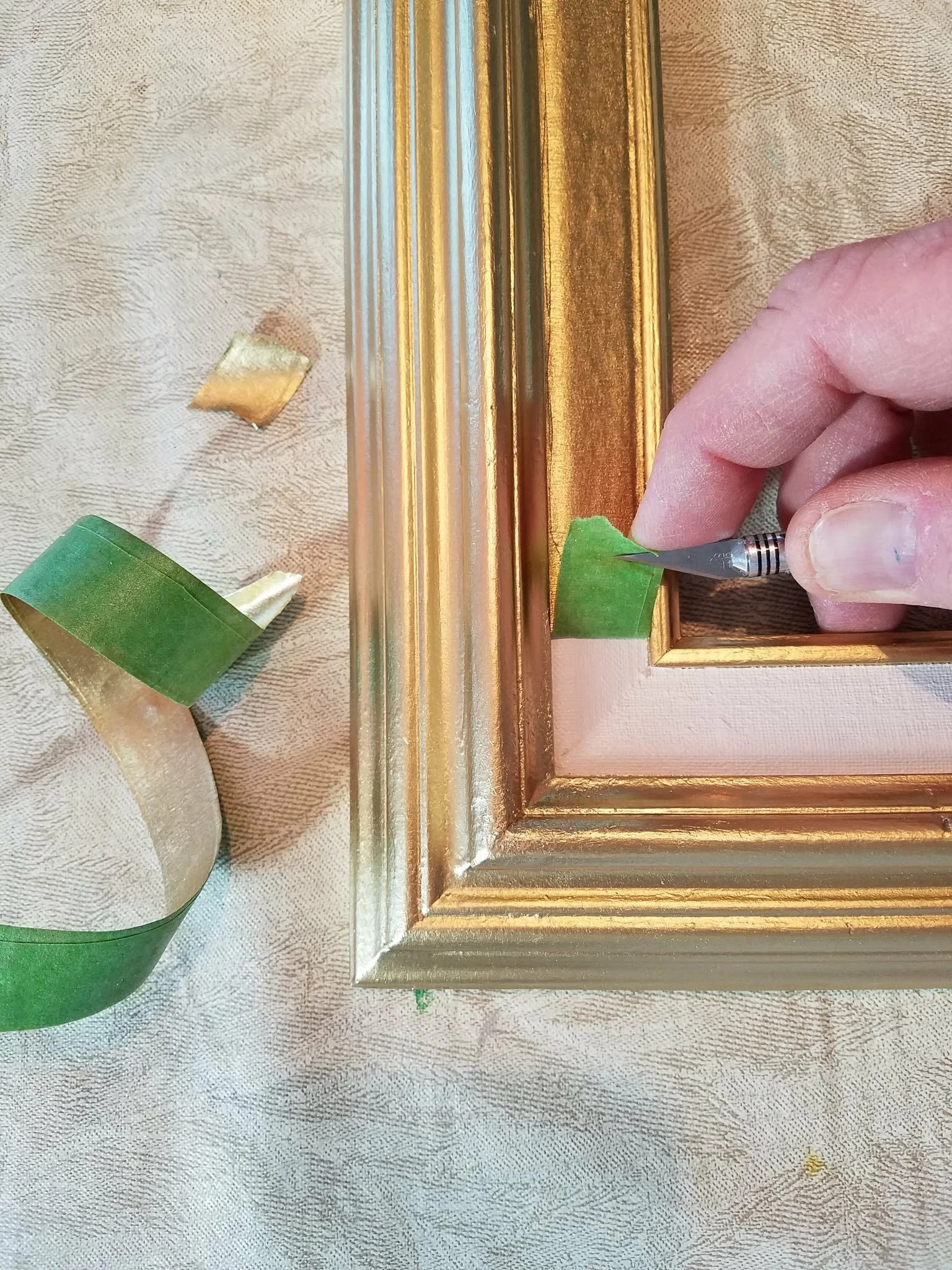 Close-up of a person painting a gold-colored picture frame with a small craft knife, with masking tape on the frame for precision.