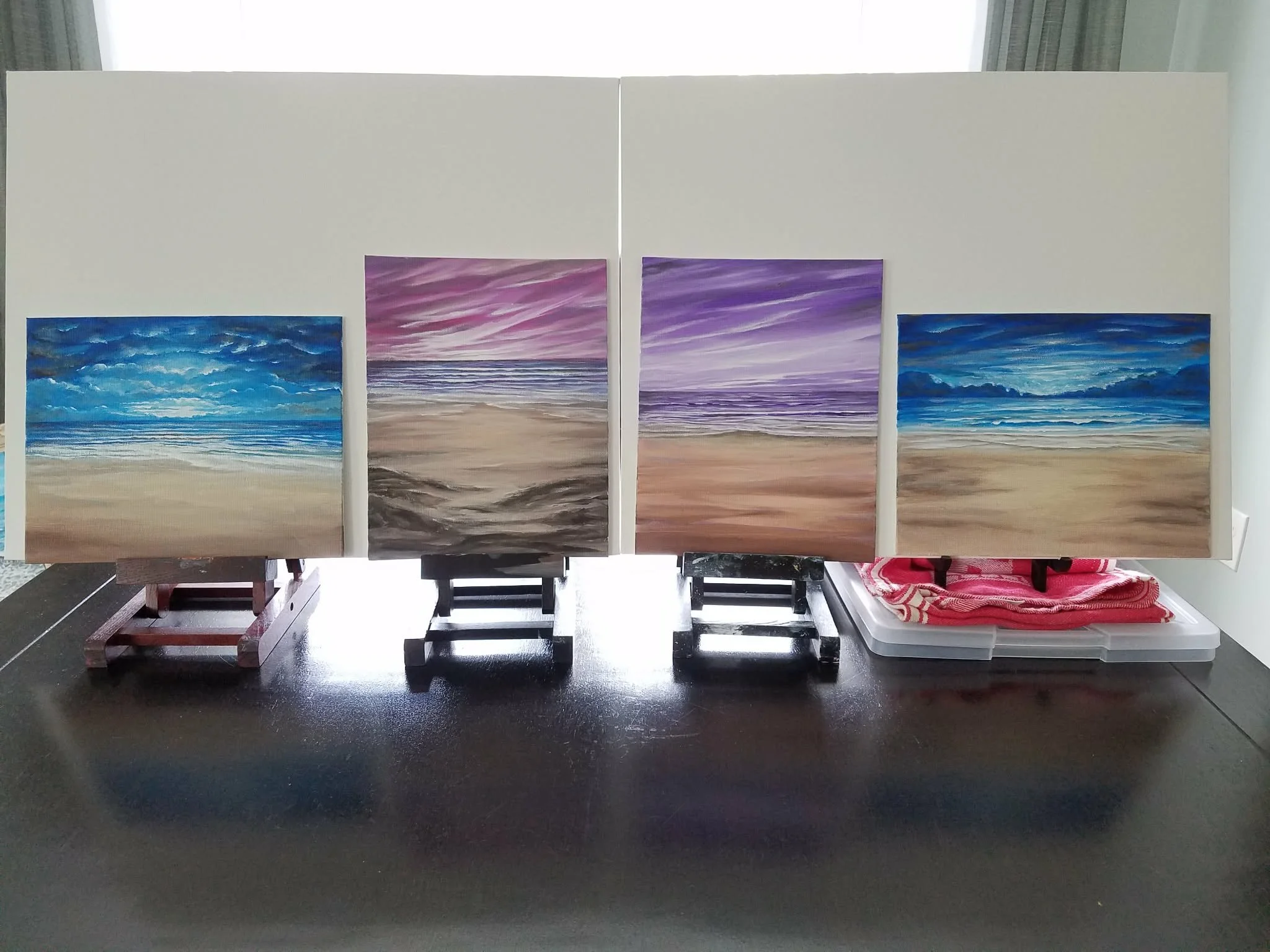 Four landscape paintings of ocean sunsets arranged on easels in front of a white wall, with sunlight shining through curtains in the background.