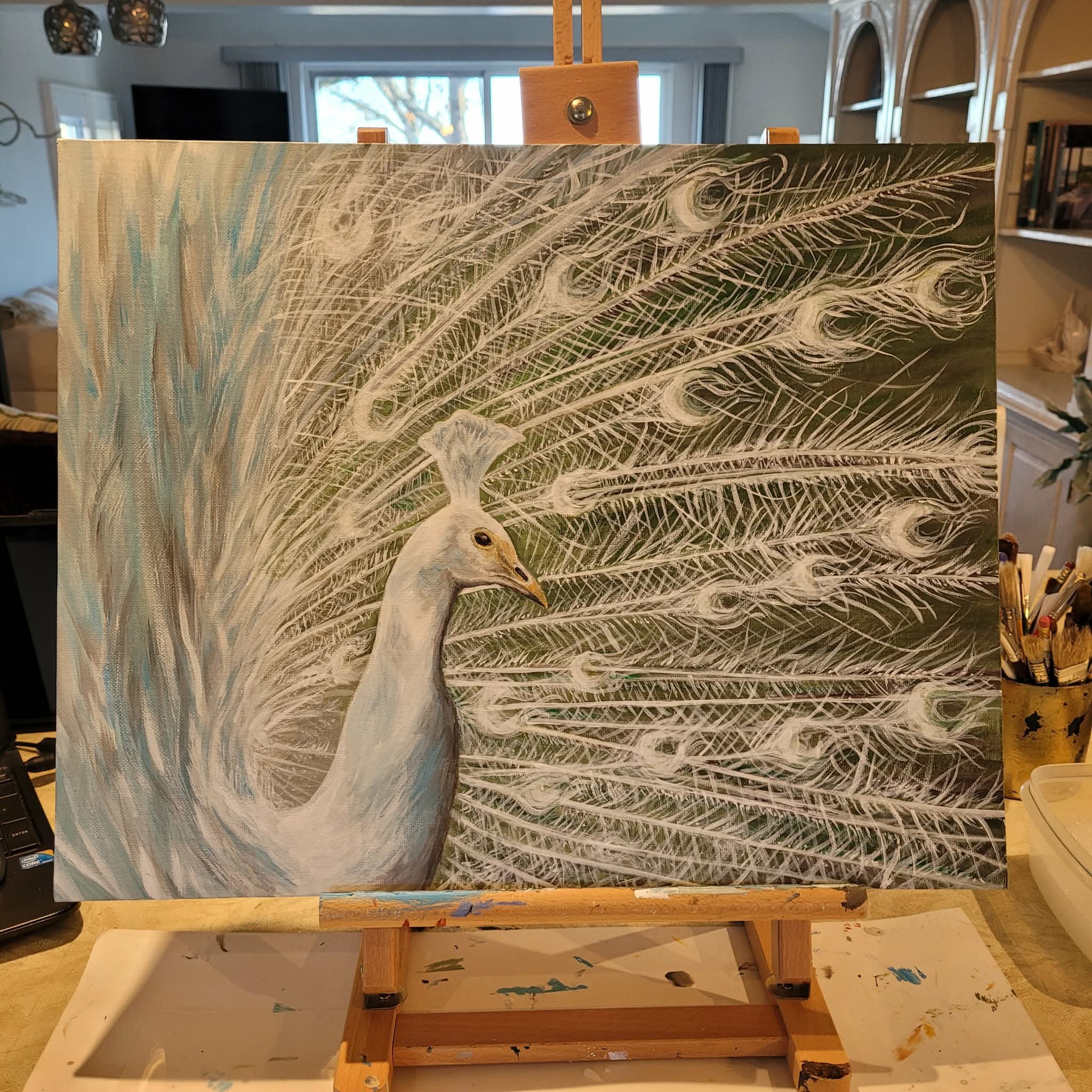 A painting on an easel features a detailed feather with eye-like patterns on the right side. The background is light blue with hints of other colors, and the canvas has visible brushstrokes and texture.