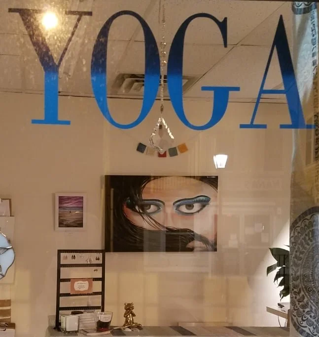 A window reflecting the word 'YOGA' in large blue letters, with indoor decor including a painting of a woman's eyes, a small framed picture, a chalkboard, and a table with a jewelry stand and decorative items visible inside.