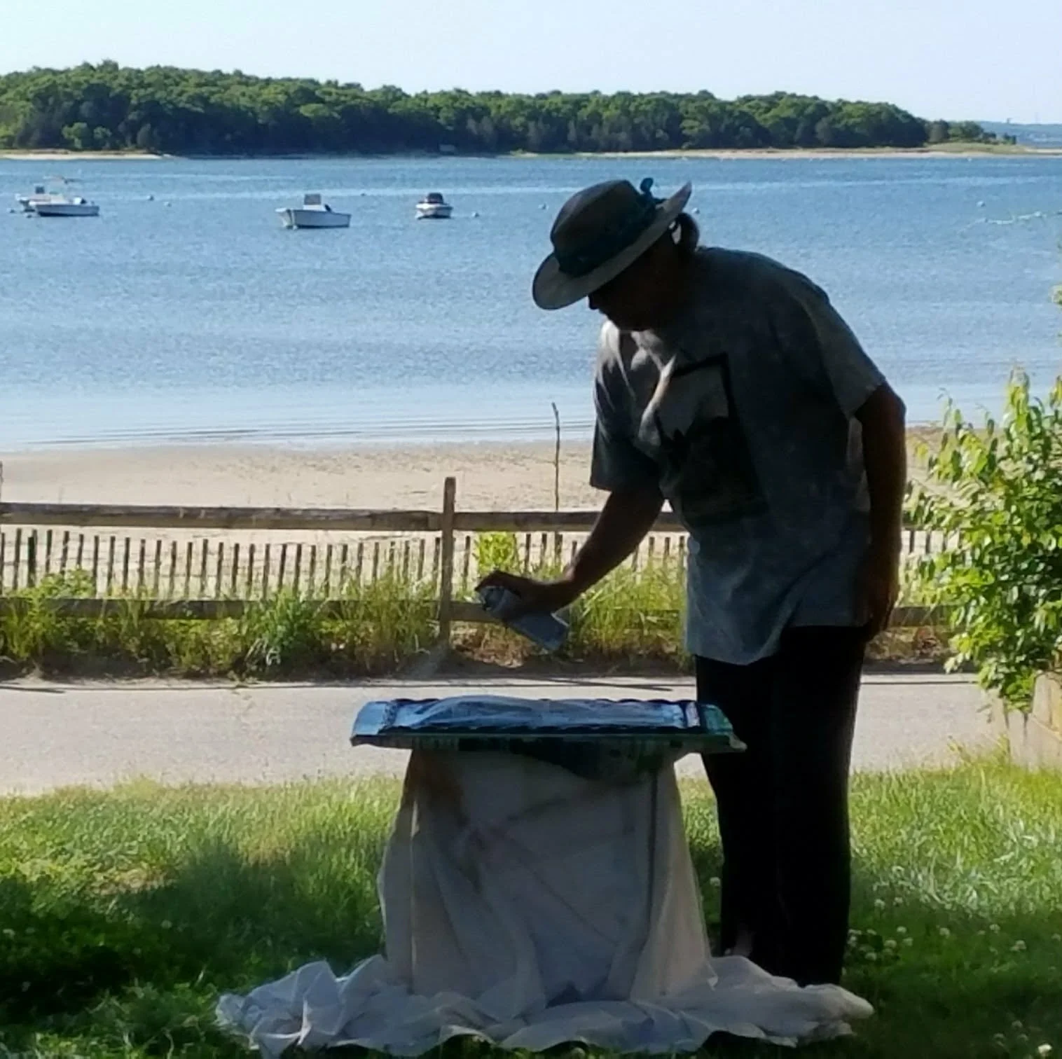 A man wearing a hat and outdoor clothing standing next to a table, looking down at a paper or book in his hand, with a lake and boats in the background.
