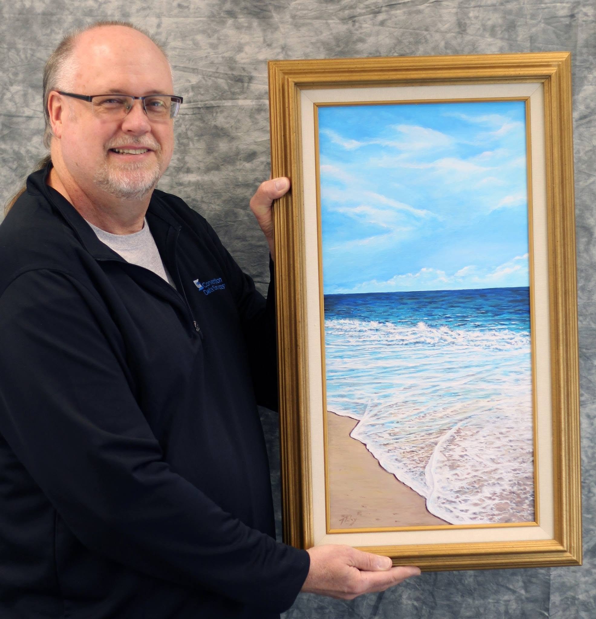 A man with glasses smiling while holding a framed landscape painting of a beach with blue sky, clouds, and ocean waves.