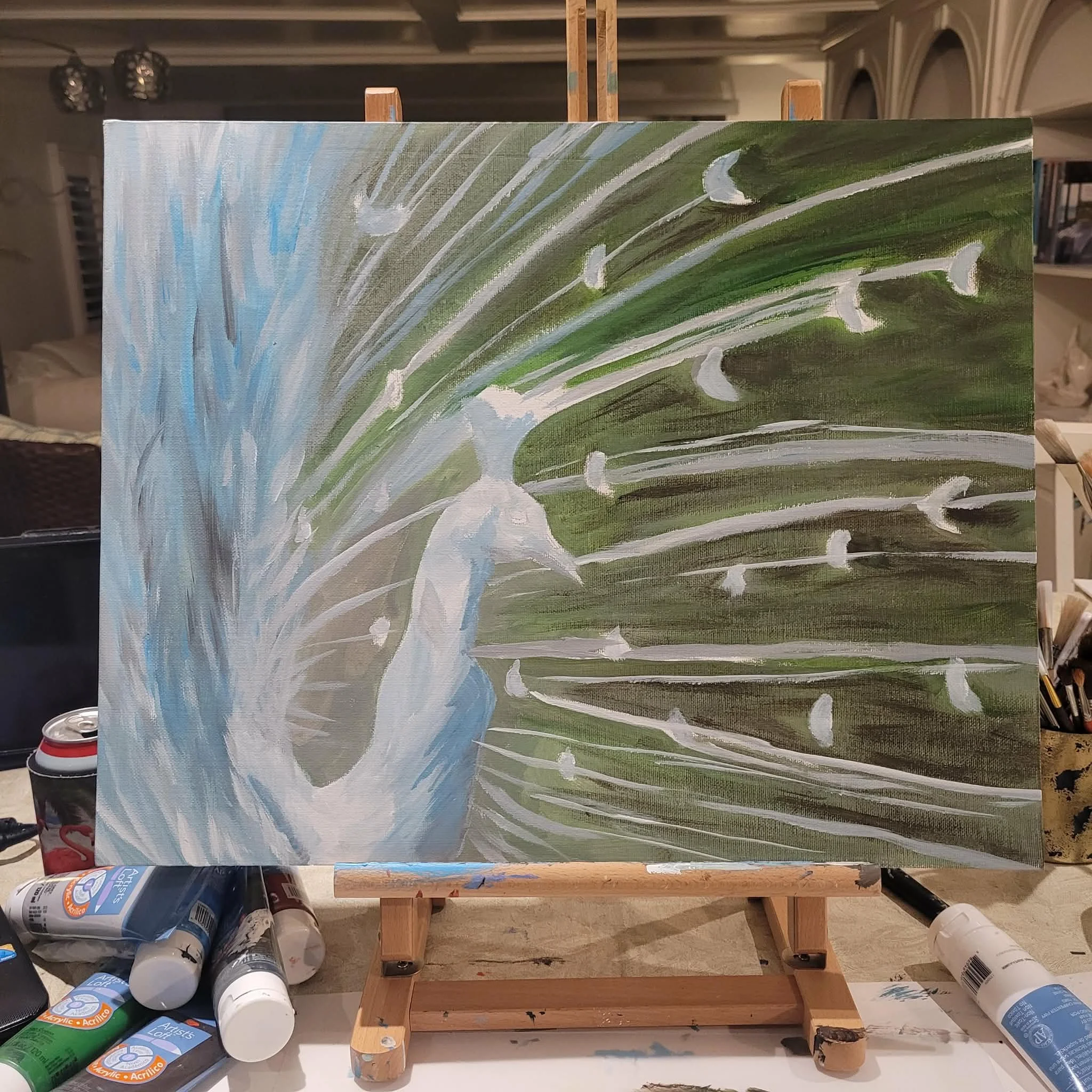 A partially painted abstract landscape on canvas on an easel, with paint bottles scattered around on a table.