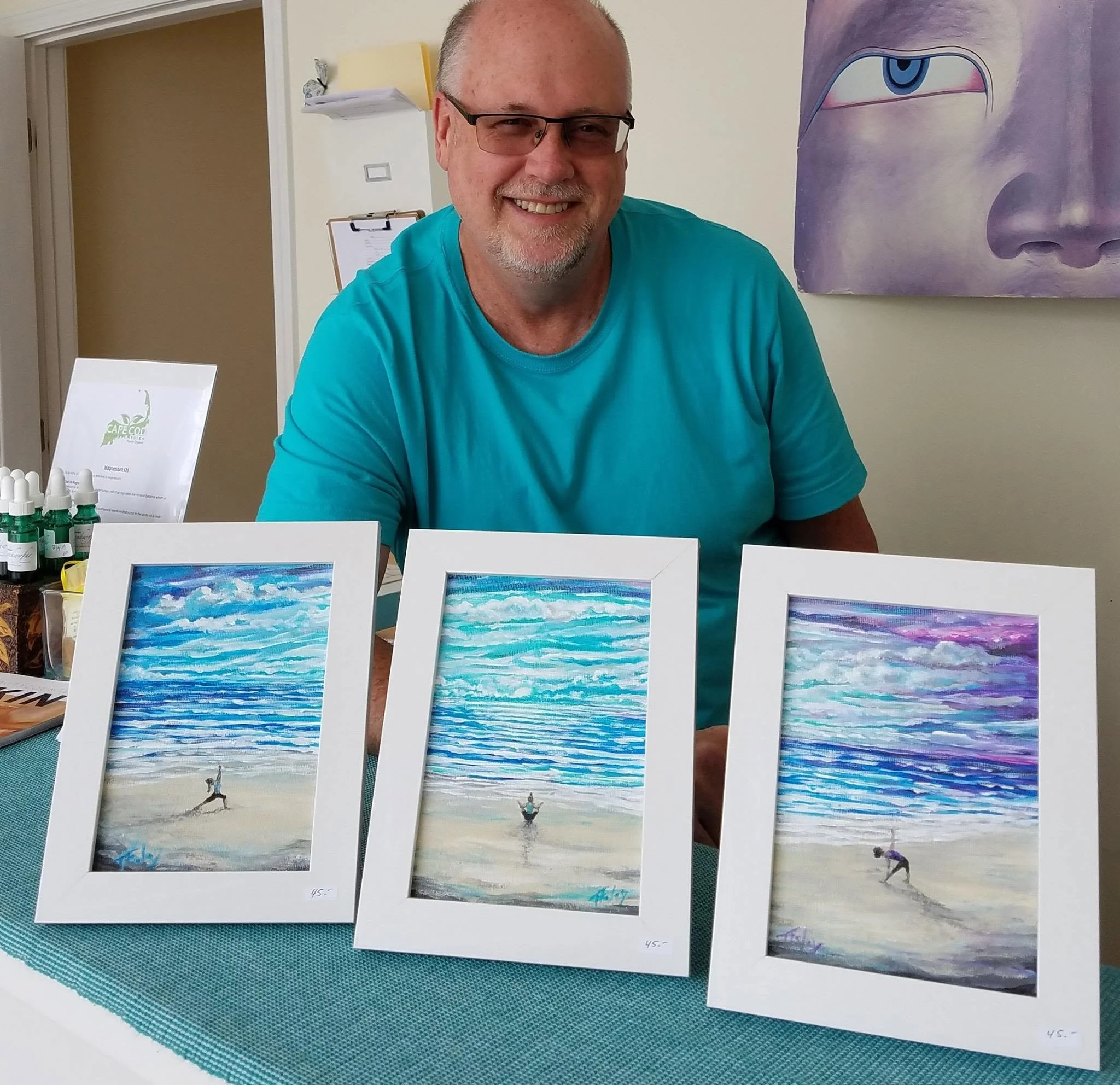 Display of four framed watercolor paintings of ocean scenes, each with clouds and waves, on a table with a teal placemat at an art show or gallery.