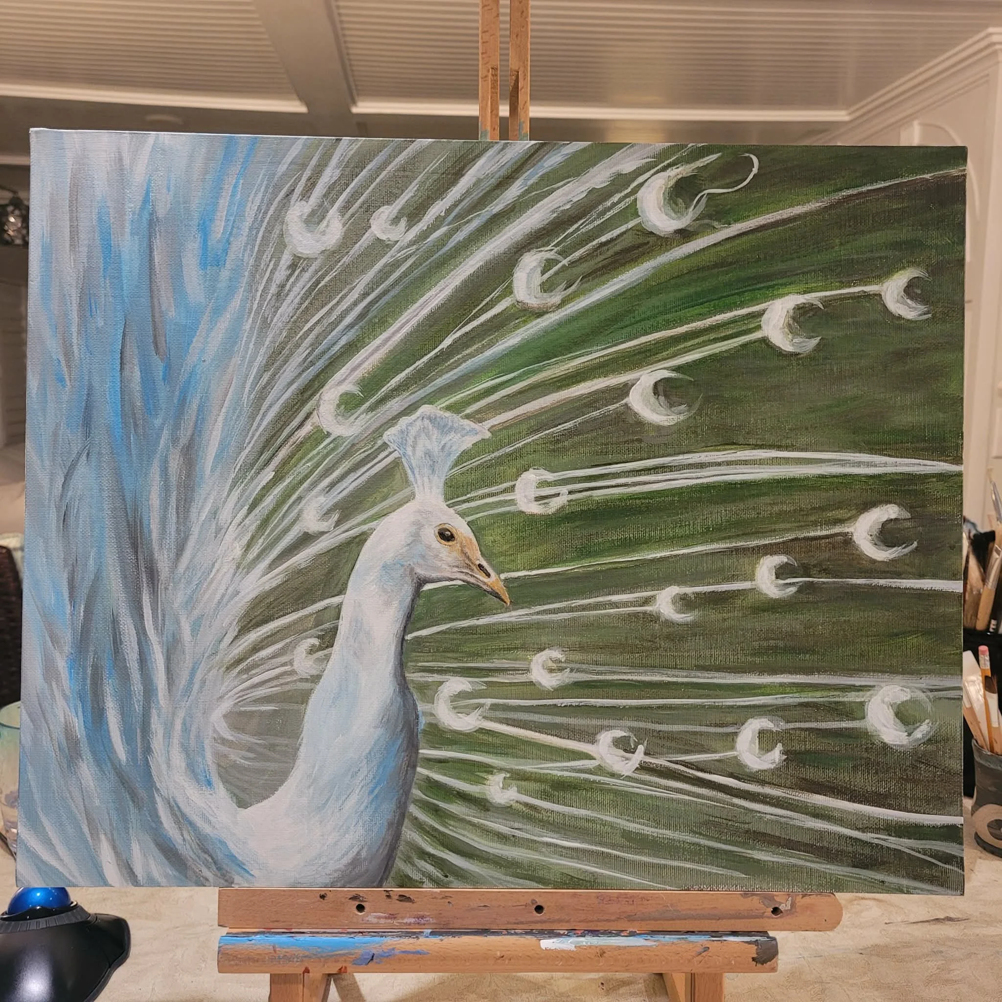 A painting of a peacock featuring blue, green, and white colors on an easel in an indoor setting.