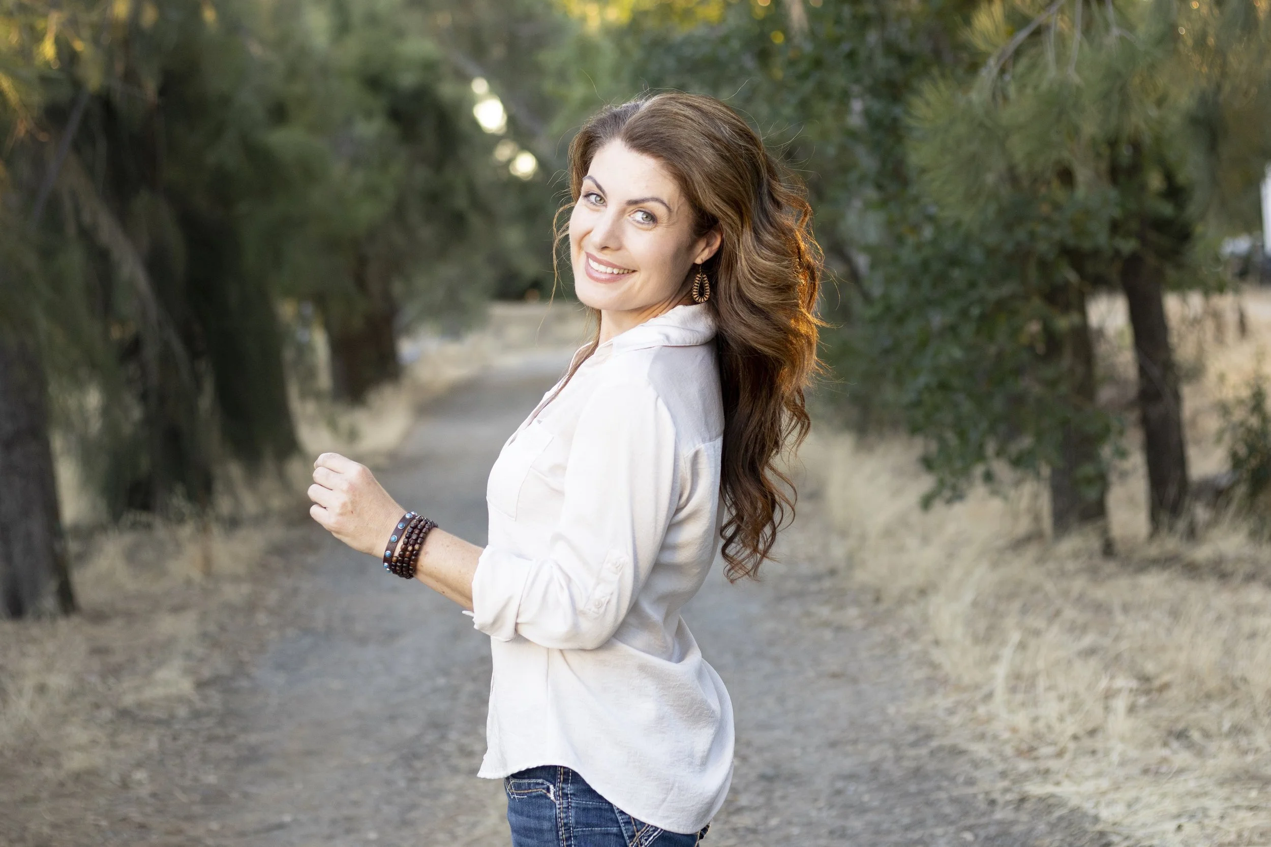 A woman with long wavy brown hair and blue eyes smiling and looking back at the camera while standing outdoors on a dirt path surrounded by trees and foliage.