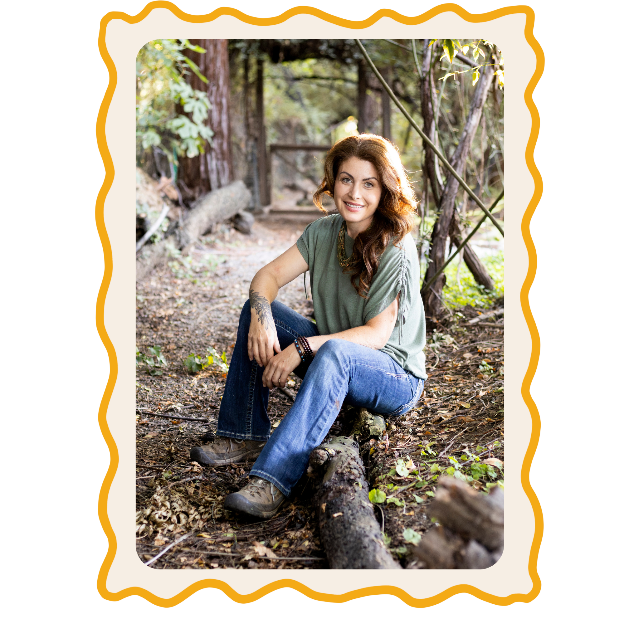 A woman with red hair and tattoos on her arm sitting on a log in a wooded area smiling at the camera.