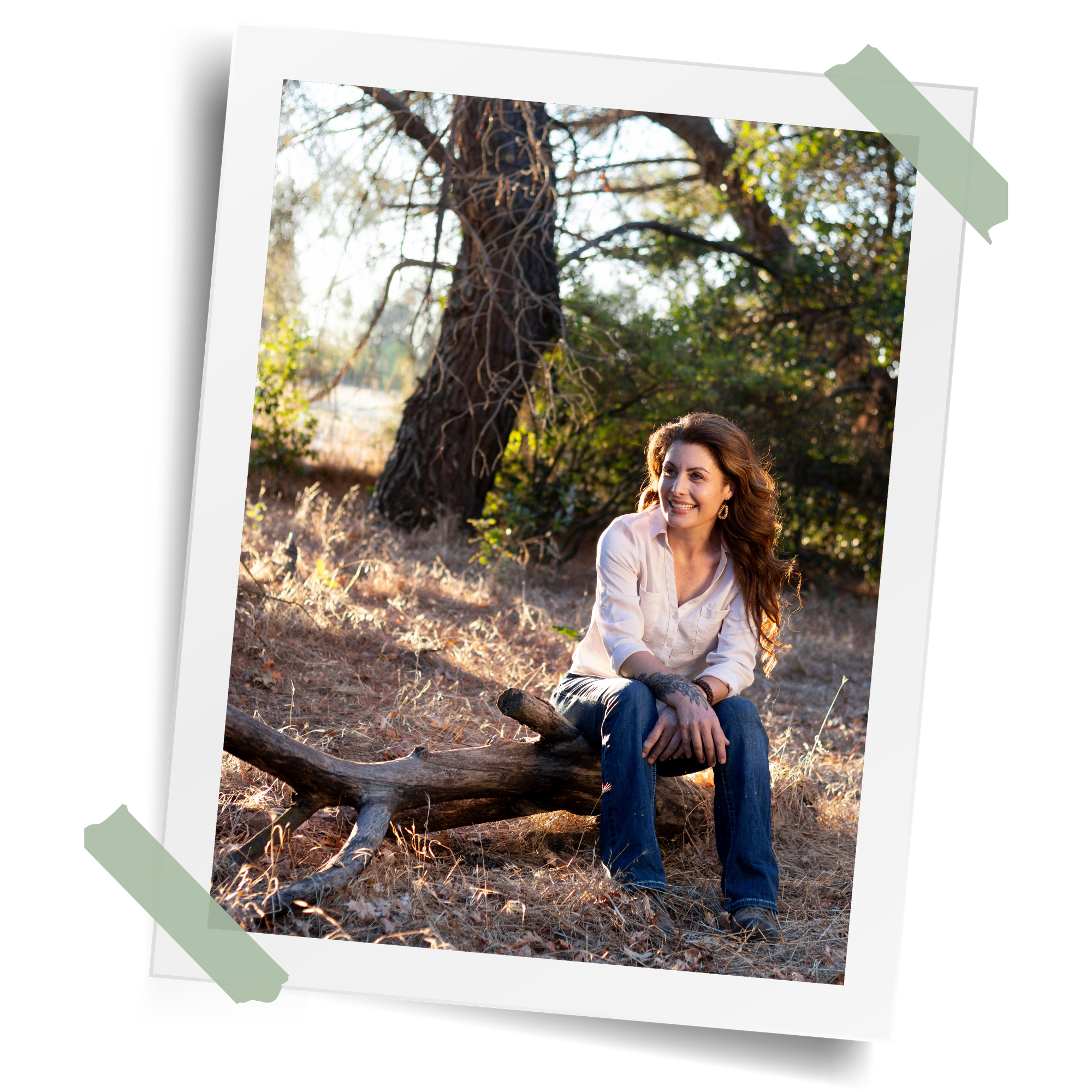 A woman with long red hair smiling and sitting outdoors near a large tree with sunlight filtering through the branches.
