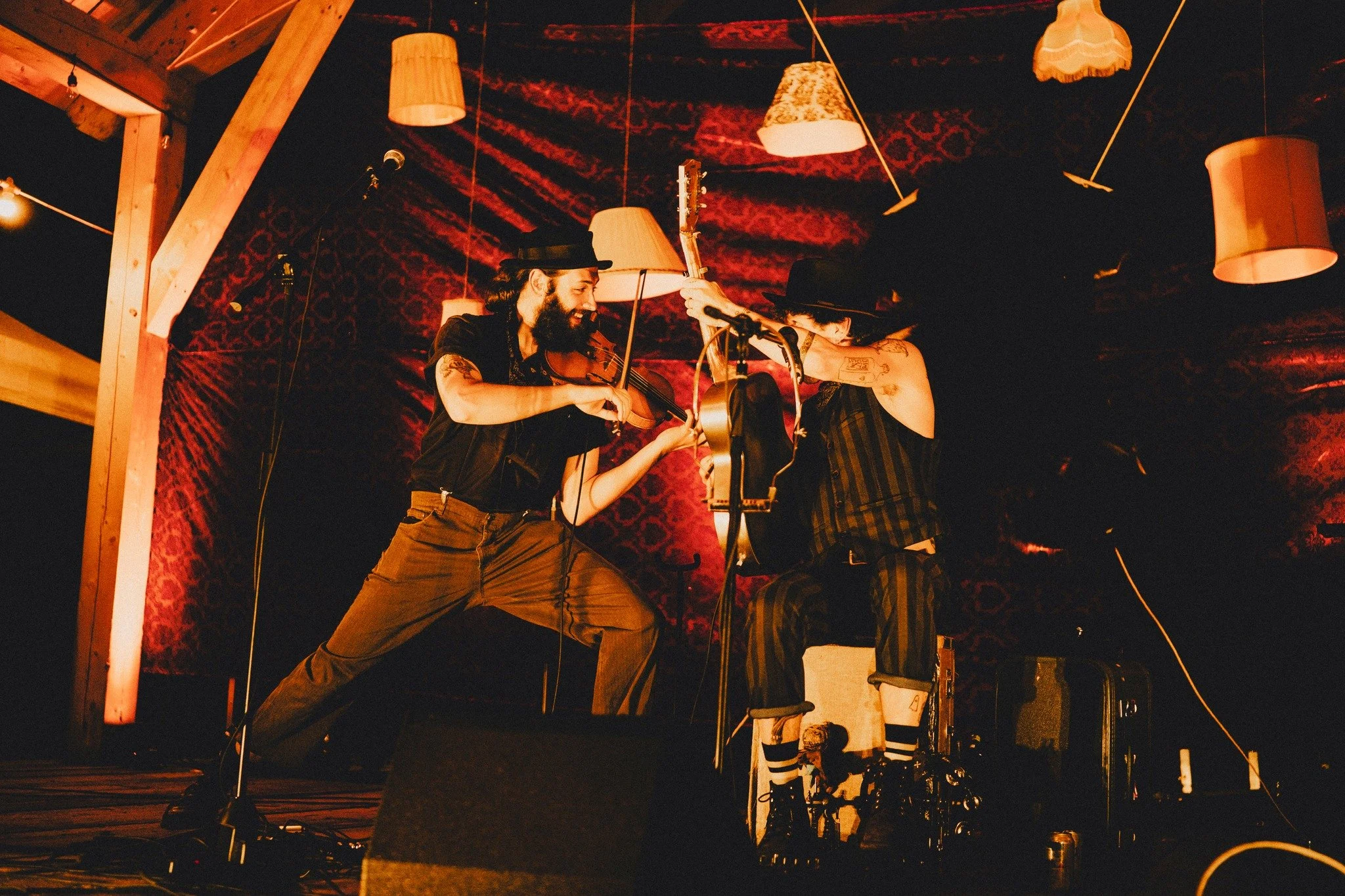 Two male musicians performing on a stage with warm lighting, one playing a violin and the other playing the guitar.