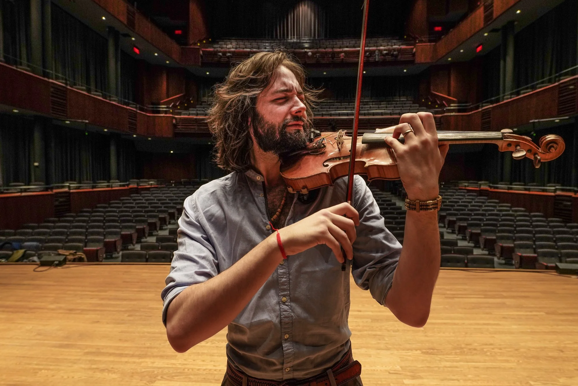 A man with long hair and a beard playing the violin on a stage inside a concert hall. The hall has multiple levels of seating with black chairs, and the stage is empty aside from the man and the violin.