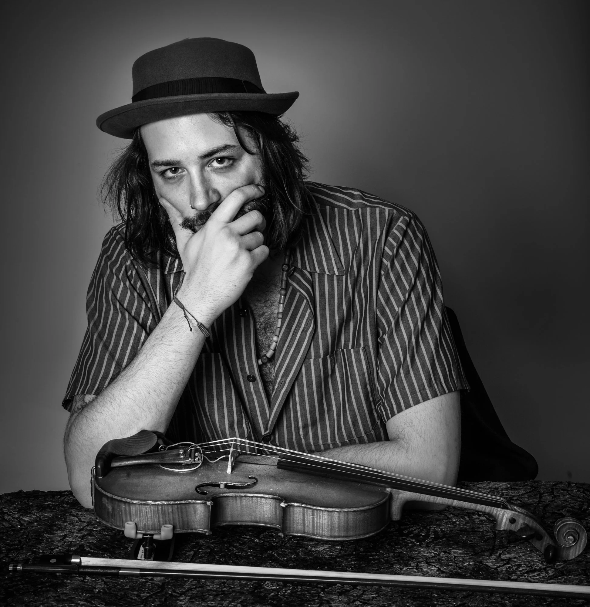 Black and white portrait of a man with long hair, wearing a wide-brimmed hat and striped shirt, resting his face on his hand with a violin on a table in front of him.