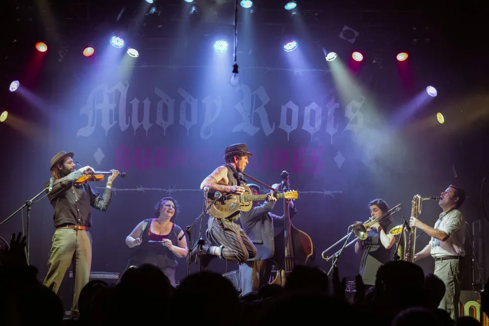 Musicians performing on stage with a backdrop that reads "Muddy Roots Argentina." One plays the violin, another the guitar, and others play a double bass, trombone, and saxophone.