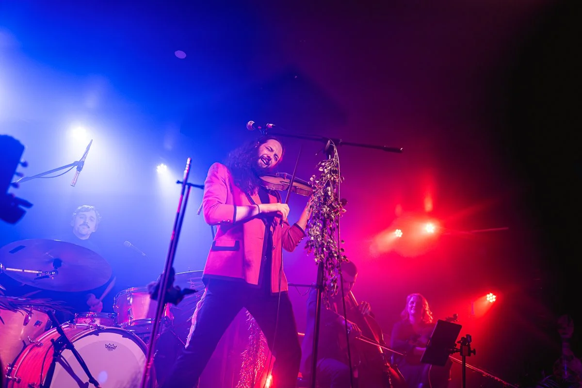Musicians performing on stage with colorful lighting, featuring a violinist dressed in red playing passionately, accompanied by a drummer and other instrumentalists.