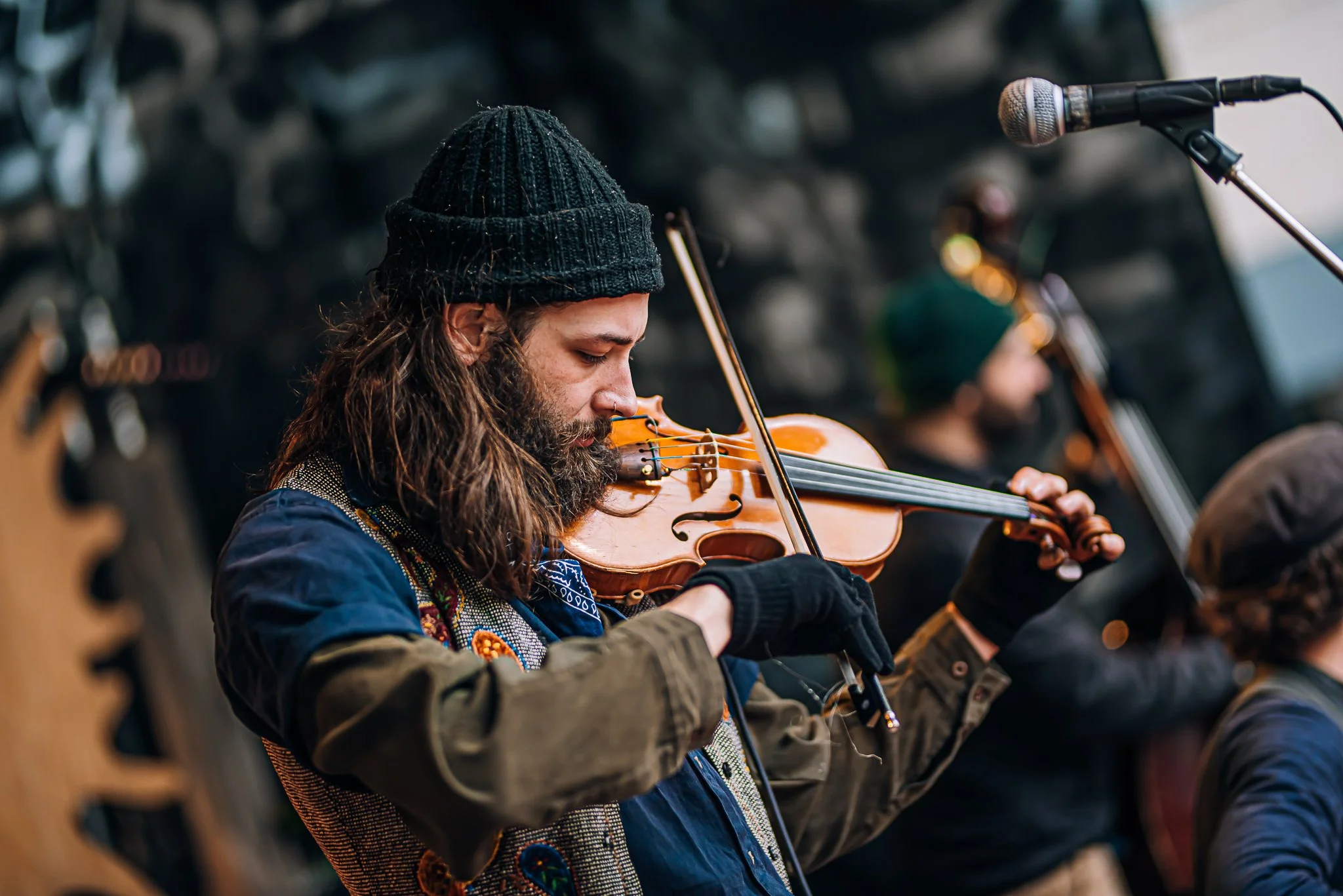 A man with long hair and a beard, wearing a black beanie and vintage clothing, tuning his violin with concentration. Microphone on stand is placed nearby. Other musicians are visible in the background.