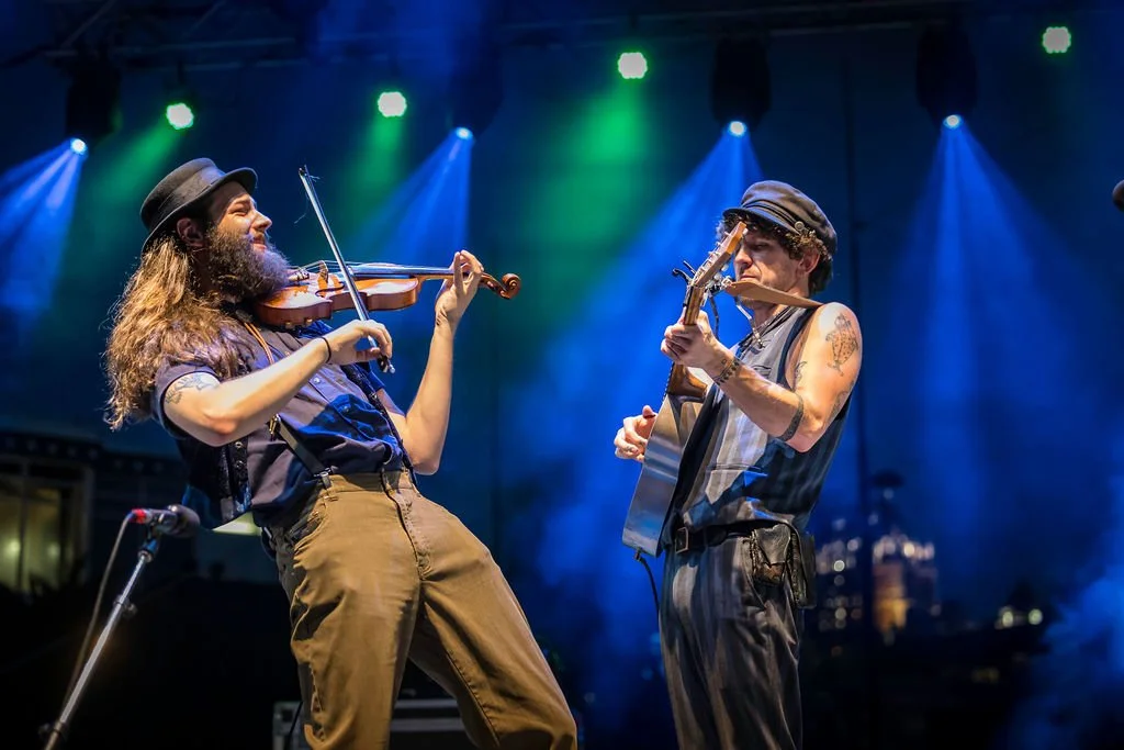 Two musicians performing on stage under blue and green lights, one playing a violin and the other a resonator guitar.