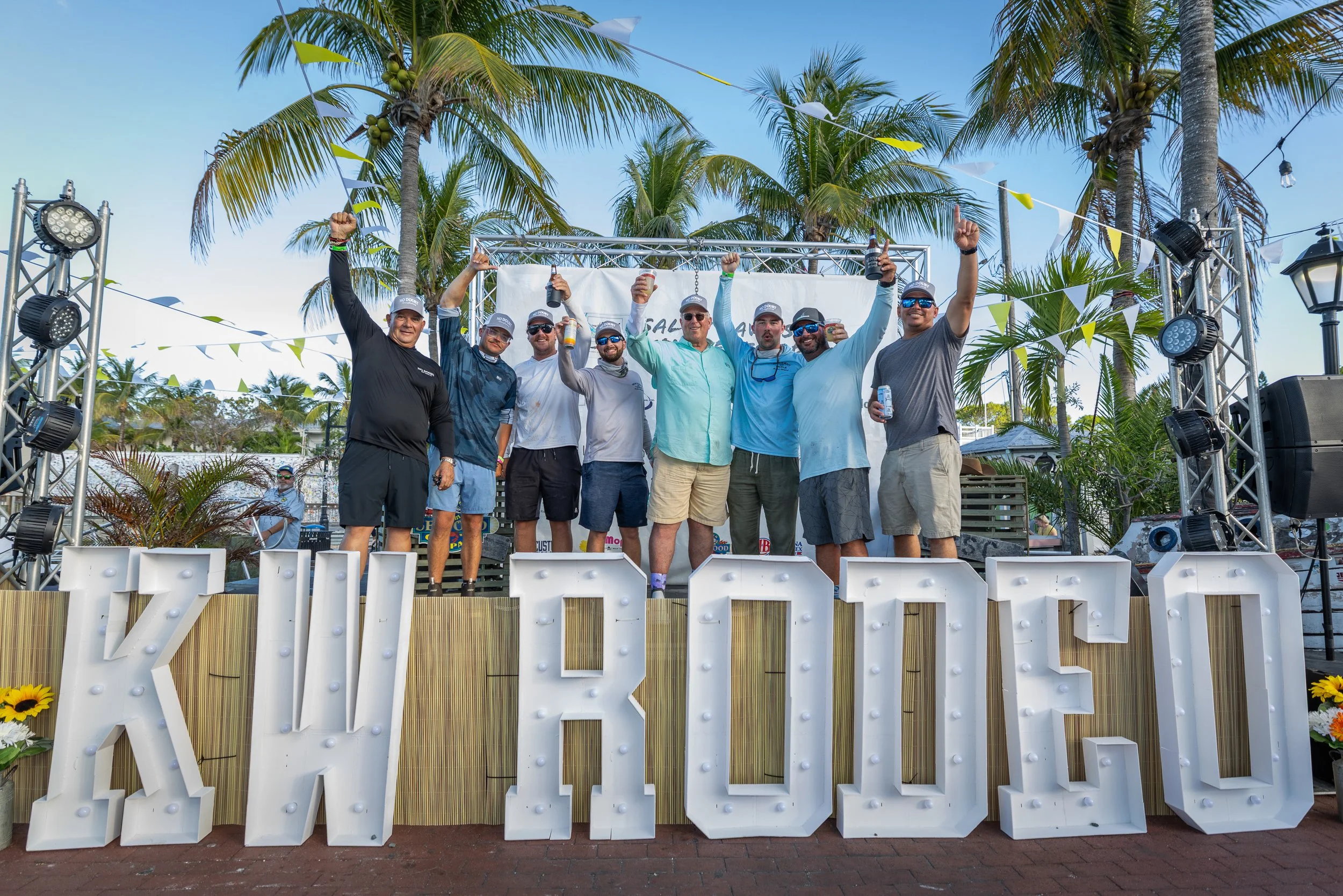 Group of men celebrating outdoors under palm trees, holding drinks, on a decorated stage with a large illuminated sign reading 'RELAX'.