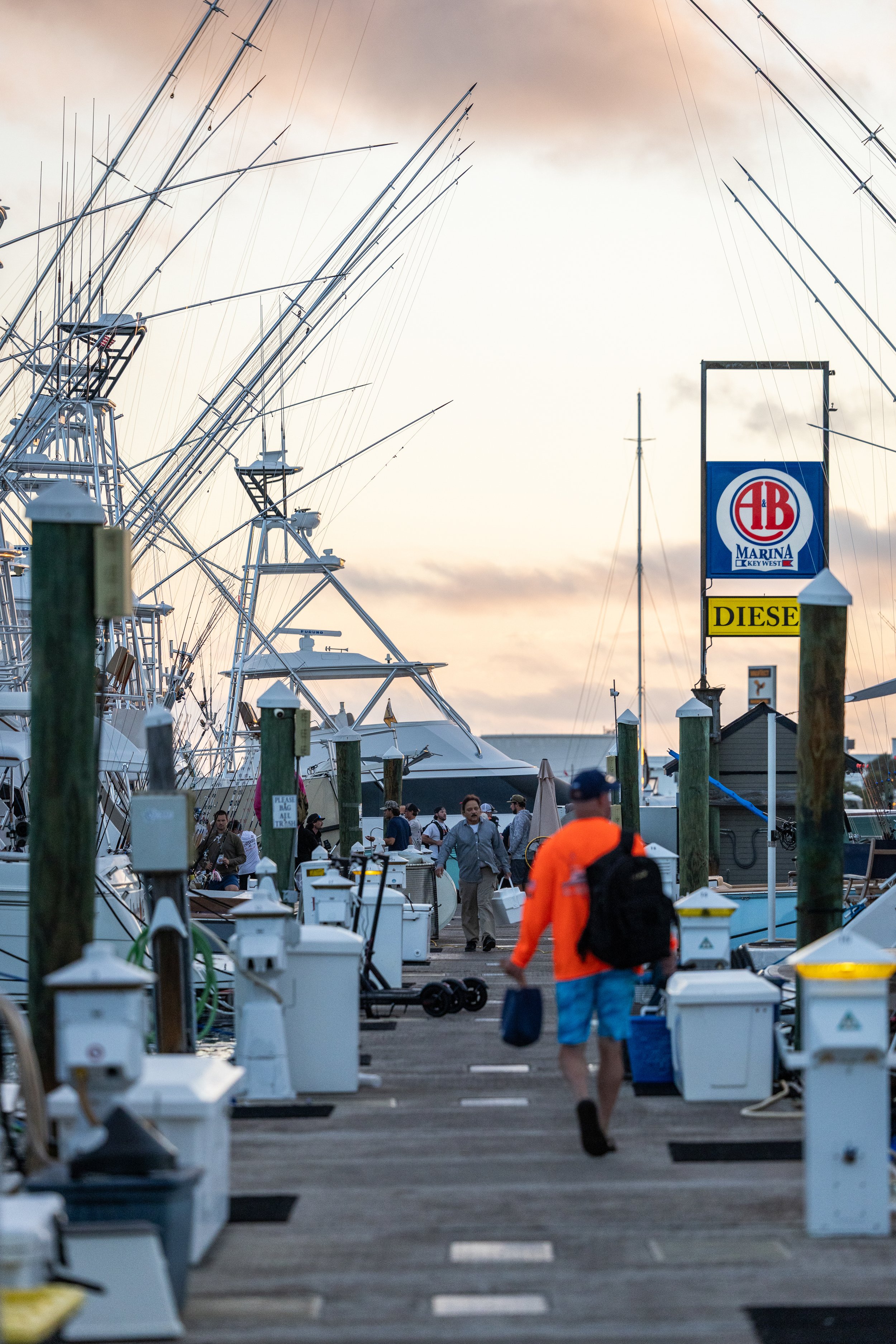 People walking along a dock filled with boats and yachts, with a clear sky and some clouds in the background, and a sign reading 'DIESEL' on the right.