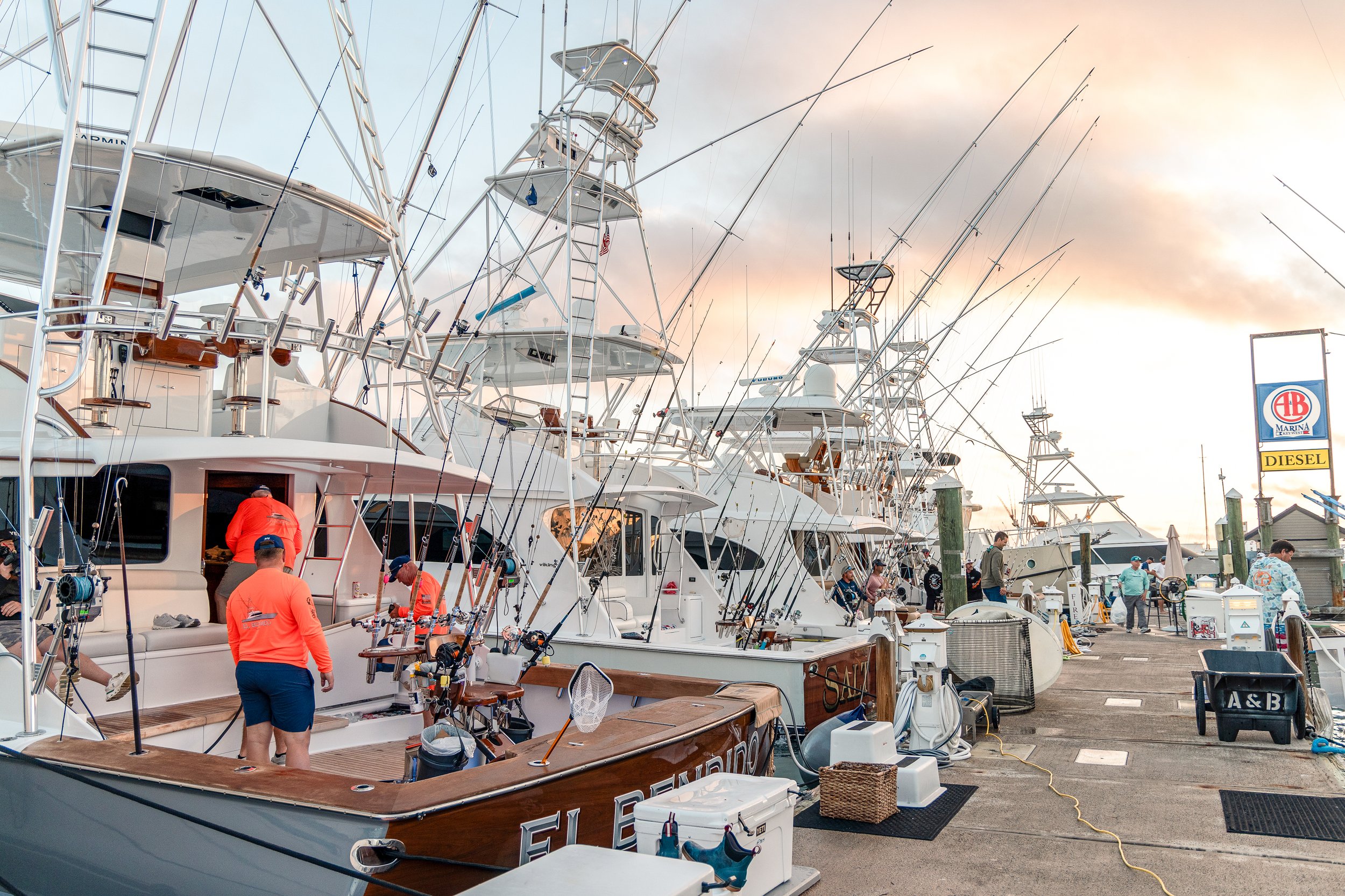 Marina with multiple sailboats docked, people working on boats, and a sign that reads "AB Marina" with a yellow sponsor sign below. The sky shows a sunset or sunrise.