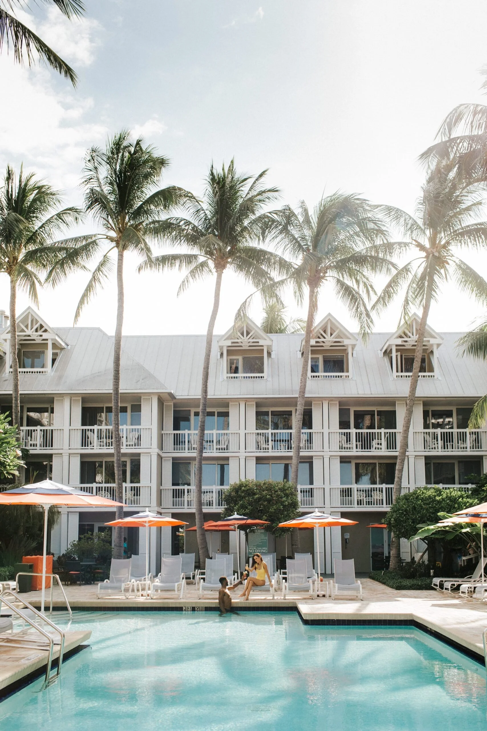 A swimming pool area in front of a multi-story hotel with white balconies, several tall palm trees, and orange umbrellas with lounge chairs underneath. A woman and a child are near the pool, one sitting on a lounge chair and the other standing in the water.