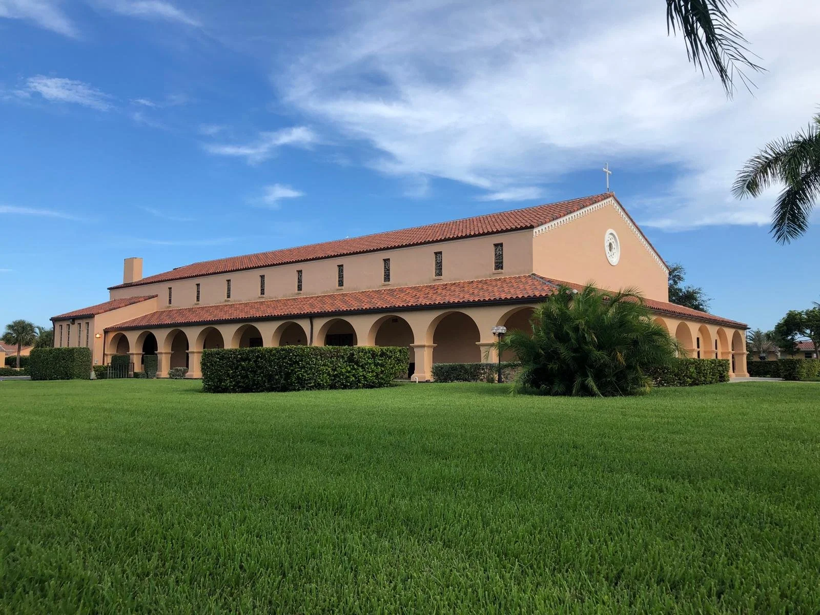 A large church with a peach-colored exterior, red tile roof, and a cross on top, surrounded by green grass, bushes, and trees under a blue sky with some clouds.