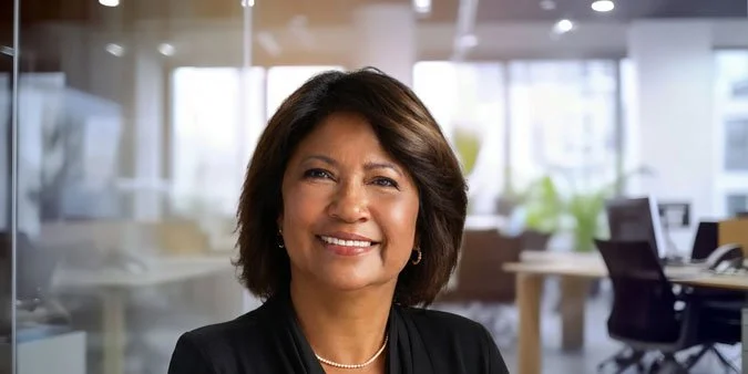 A smiling woman with shoulder-length dark hair in an office setting with large windows and desks in the background.
