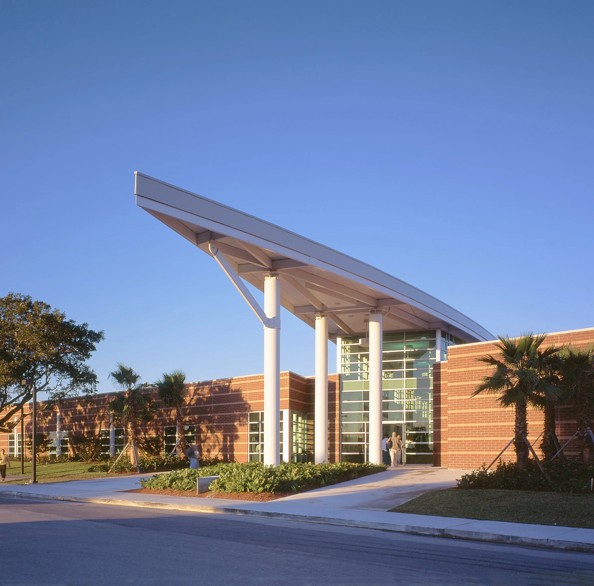 A modern building with brick and glass facade, white support columns, and a large overhang, surrounded by trees and a sidewalk on a sunny day.