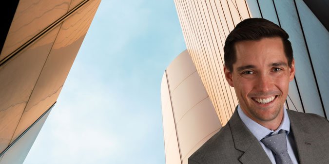 Smiling man in suit takes selfie outdoors with modern glass buildings behind him.