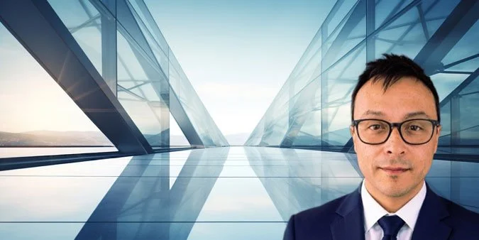 A man wearing glasses and a suit, standing in front of a modern glass building with a clear sky background.