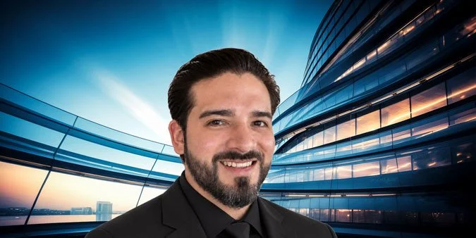 A man with dark hair and a beard, smiling, in front of a modern glass building at sunset.