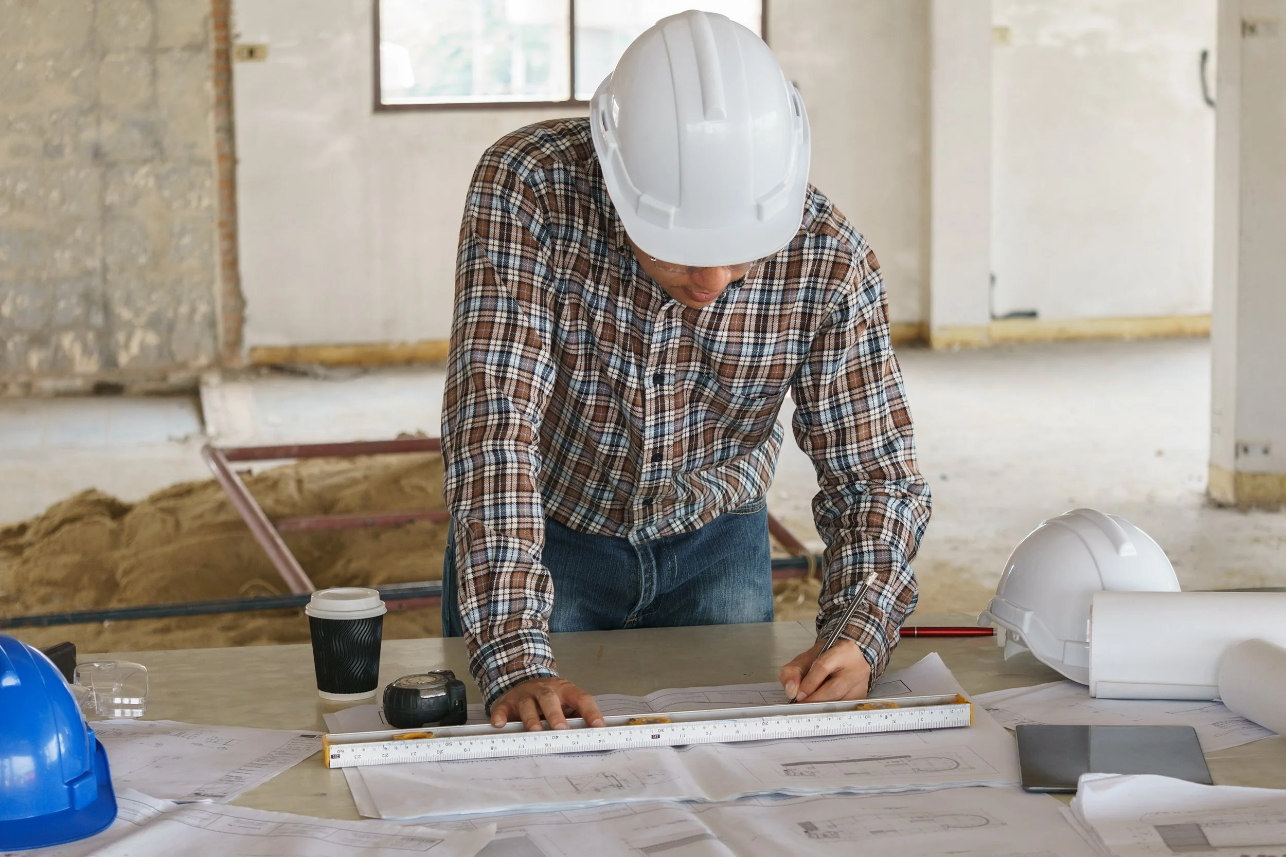 A construction worker wearing a plaid shirt and a white hard hat is working on blueprints at a construction site, with construction materials and a coffee cup on the table.