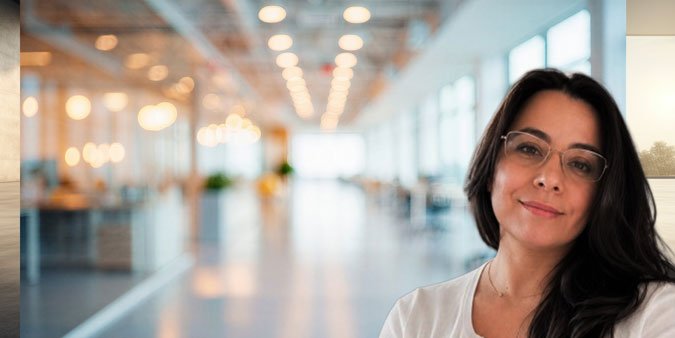 A woman with glasses and dark hair smiling in a brightly lit modern office or coworking space.