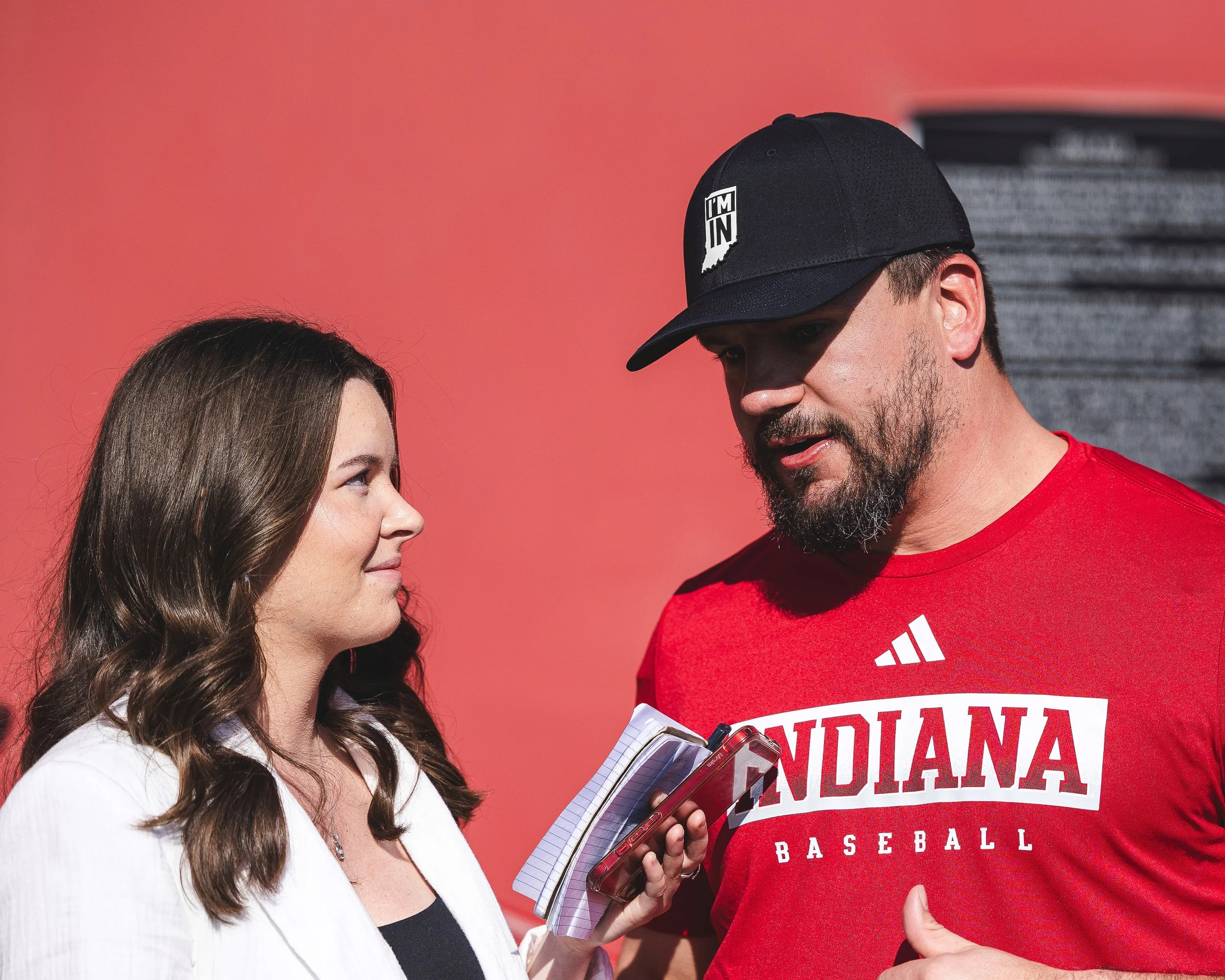 A young woman with long brown hair and a man with a beard wearing a black cap and red shirt with 'Indian Baseball' on it are having a conversation. The woman is holding a clipboard and pen, and the man is looking at her.