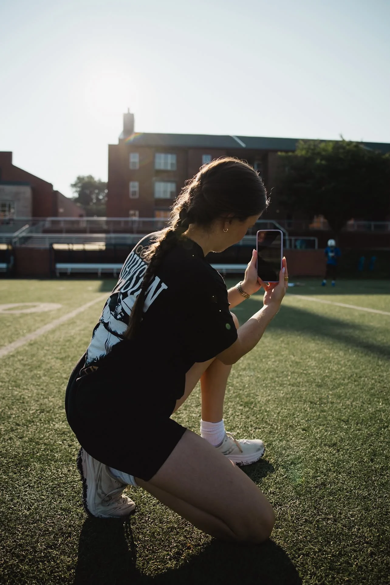 A woman with a braid kneeling on a football field, holding her phone, with a person in football gear in the background.