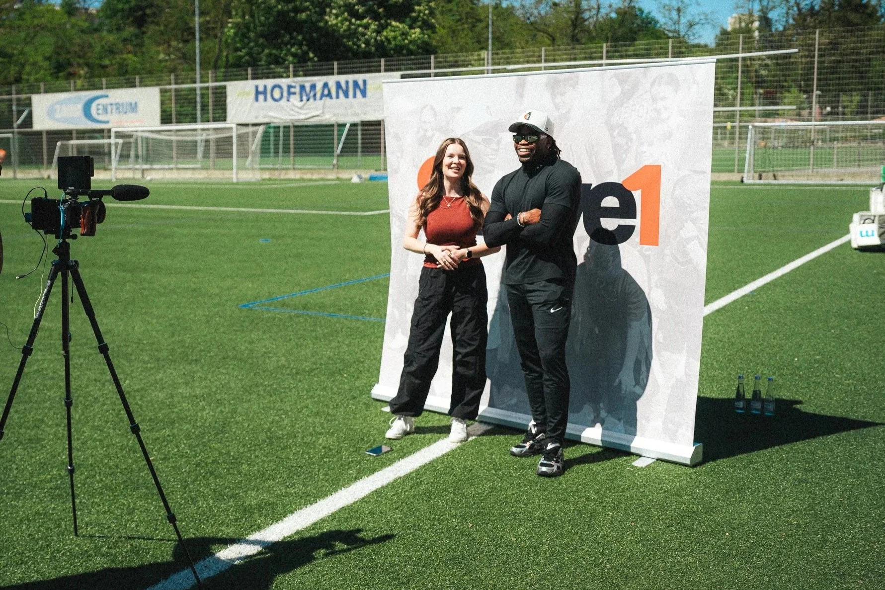 Two people standing in front of a large advertising backdrop on a sports field, with a camera on a tripod nearby. One person is a woman with long brown hair wearing a sleeveless top and black pants, smiling. The other is a man with dreadlocks, wearing a white cap, sunglasses, black long sleeve shirt, and black pants, also smiling with arms crossed. The background features a green sports field, fencing, and some advertising banners.