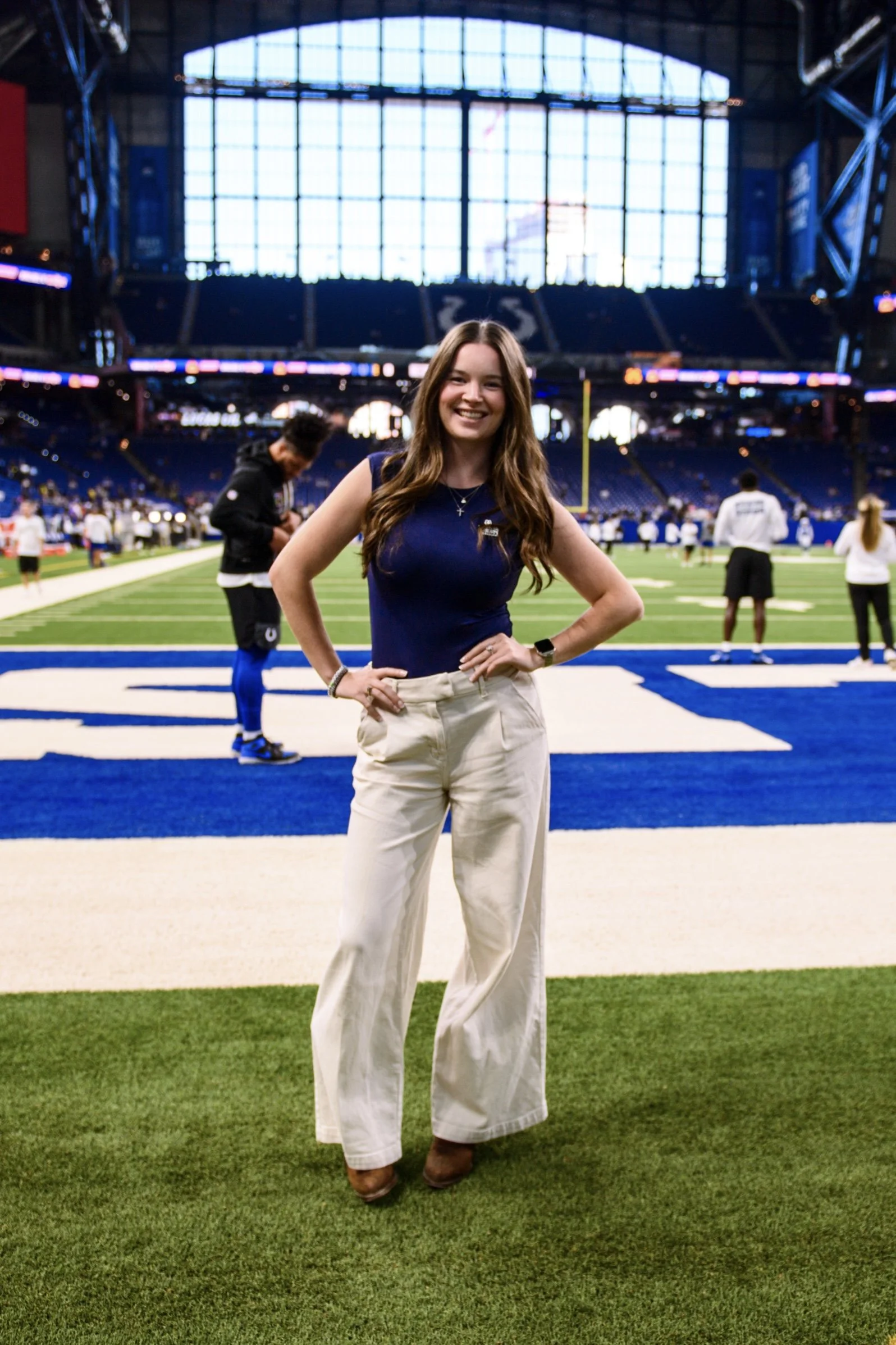 A woman standing on a football field inside a large stadium with a glass roof, smiling, wearing a navy blue sleeveless top, beige pants, and brown shoes, with other people in the background.