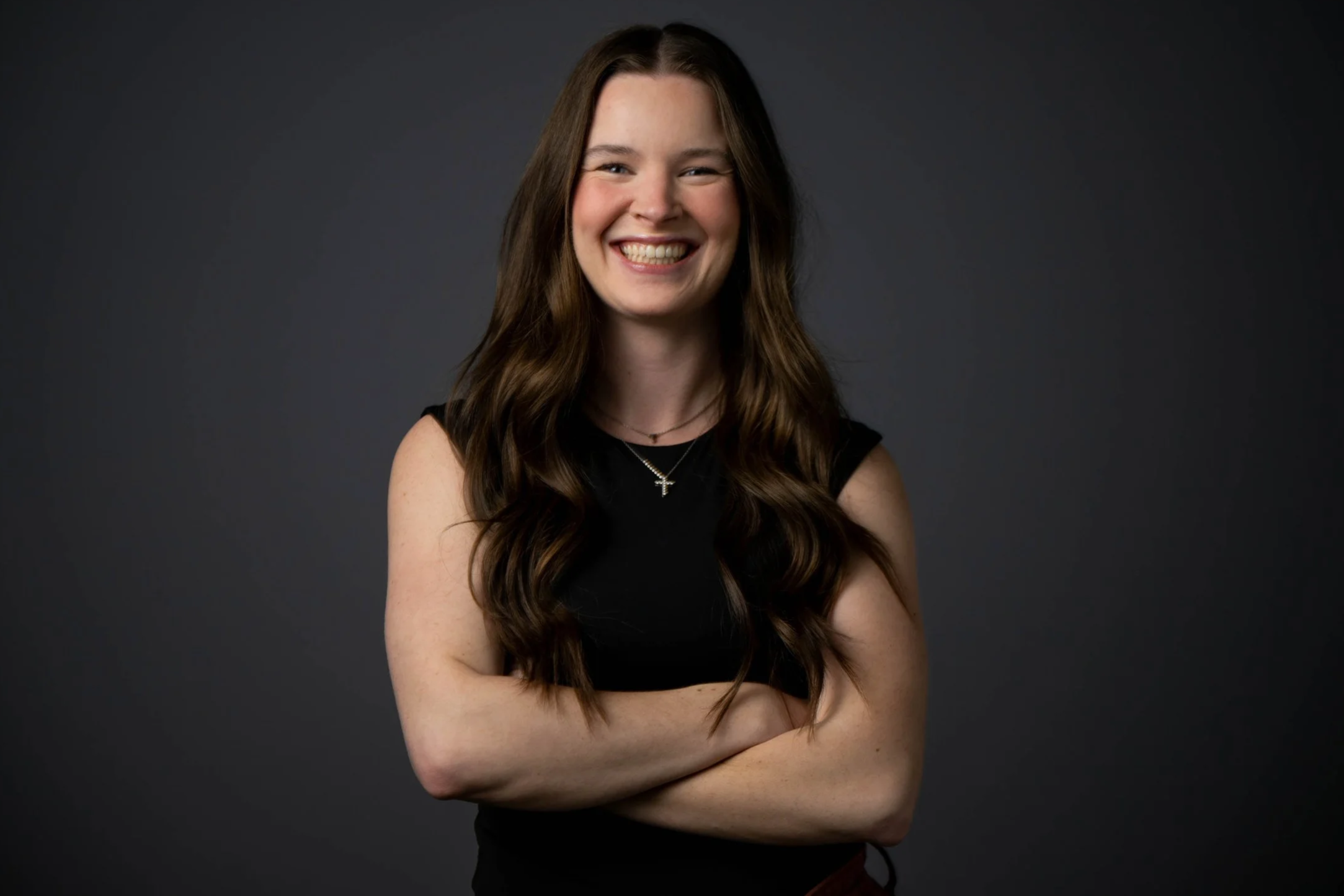 A young woman with long, wavy brown hair, smiling, with arms crossed, wearing a black sleeveless top, a cross necklace, against a dark gray background.