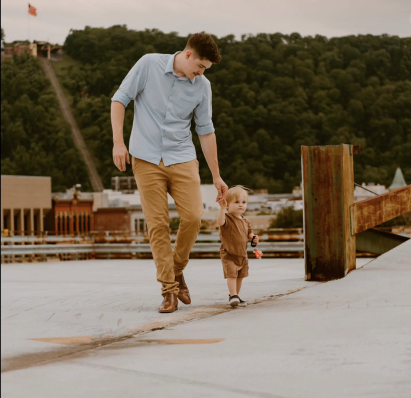 A young man holding the hand of a small child, walking together on a concrete surface outdoors with a cityscape and green hills in the background.