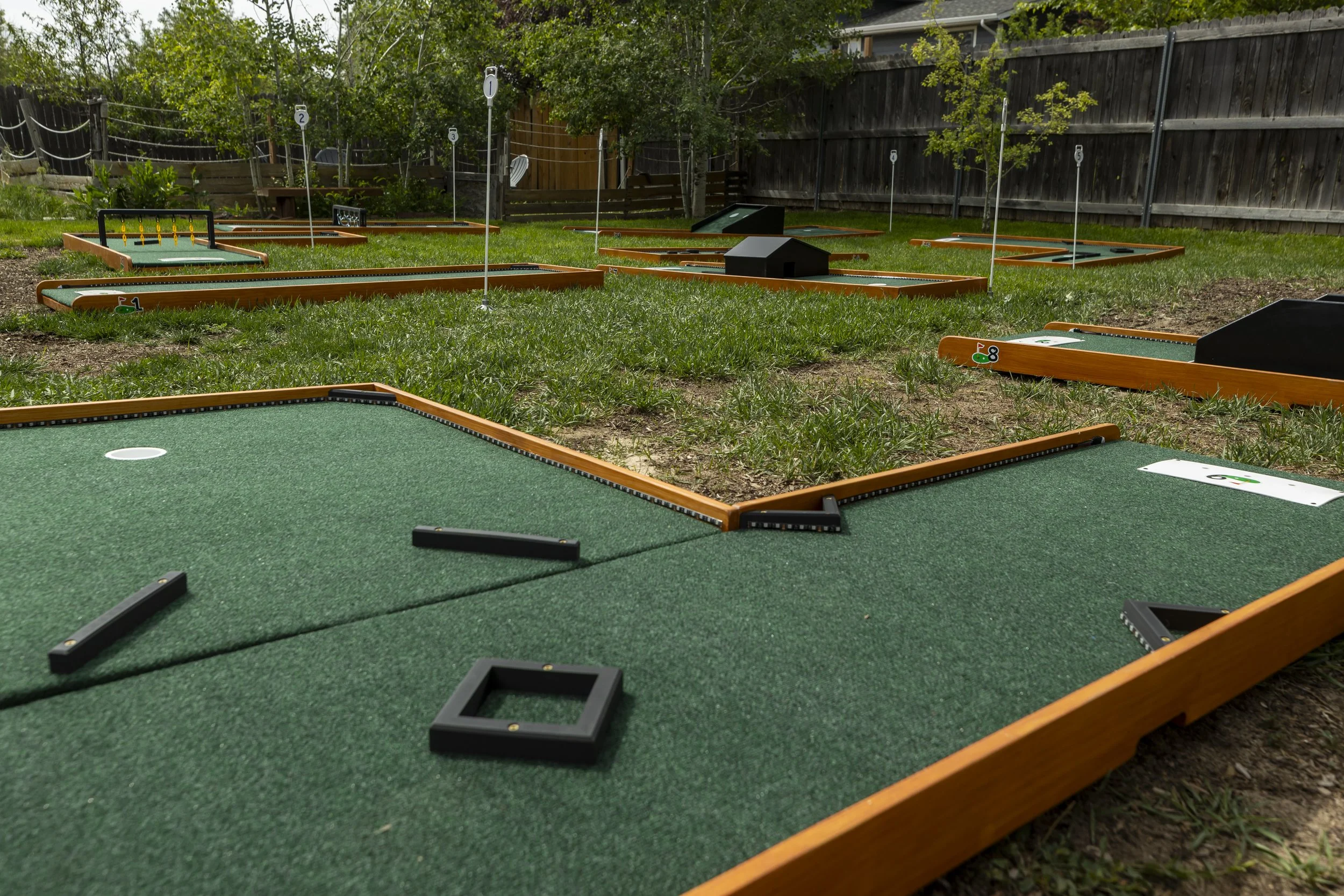 Miniature golf course outdoors with various holes marked with numbers and flags, green turf, and surrounding trees and fencing.