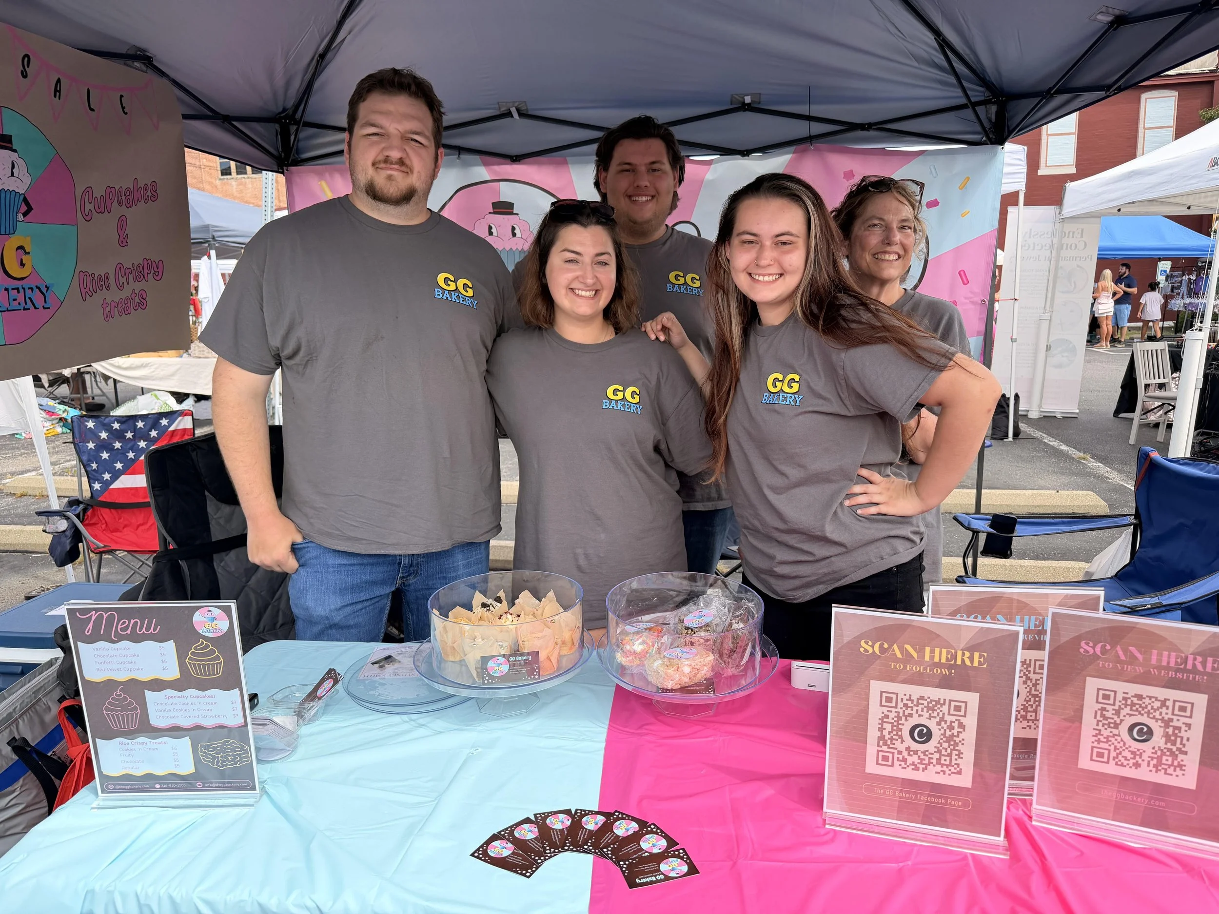 Group of five people smiling and standing behind a dessert booth at an outdoor event, with pink and blue decorations and cupcakes and treats on display.