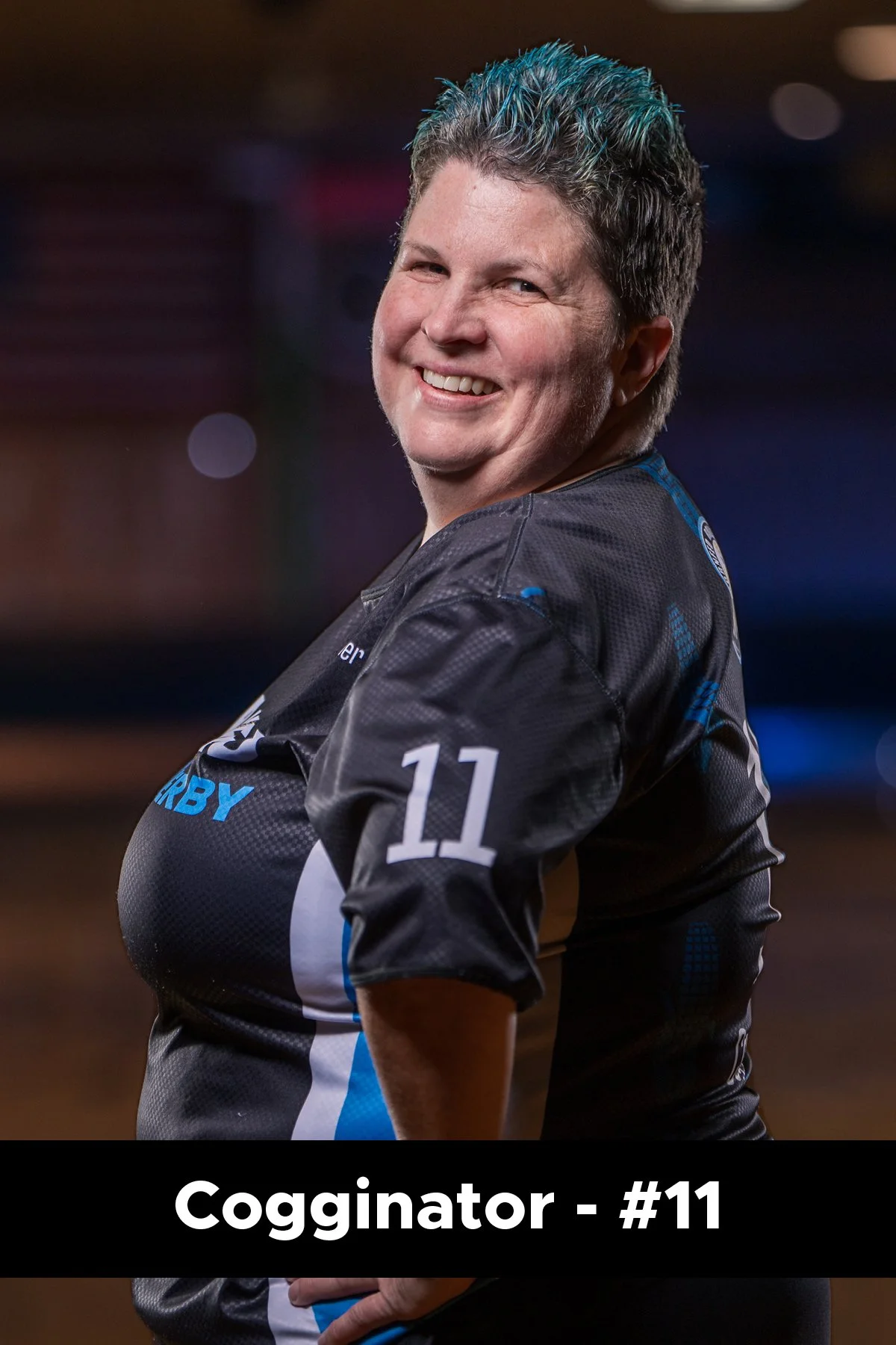 A smiling woman with short, spiky hair with blue tips, wearing a black sports jersey with the number 11 and the words 'Cogginator' on it. She has her arms crossed and is looking towards the camera.