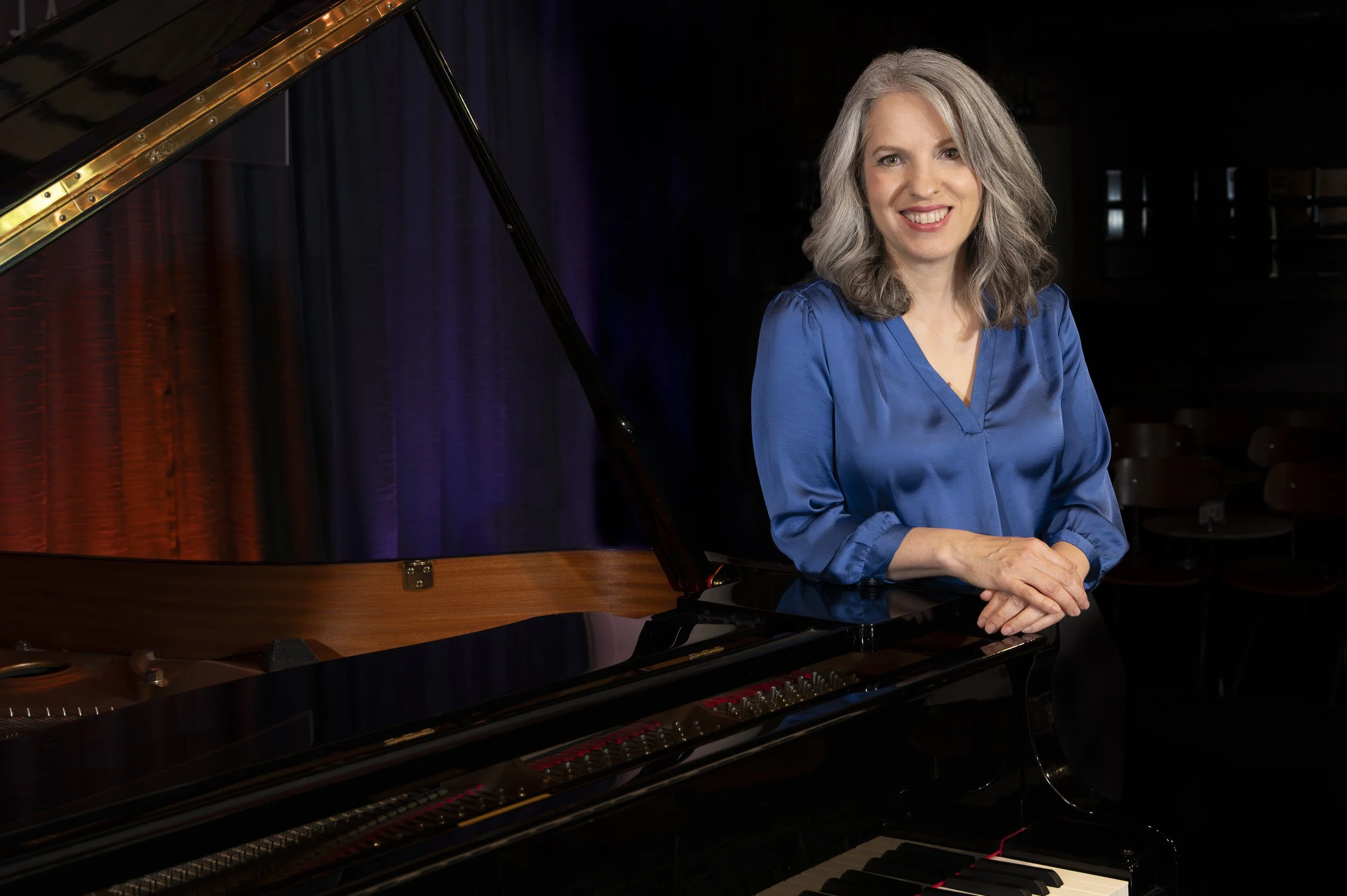 A woman with gray hair in a blue blouse smiling, seated beside a grand piano with its lid open, in a darkened stage setting.
