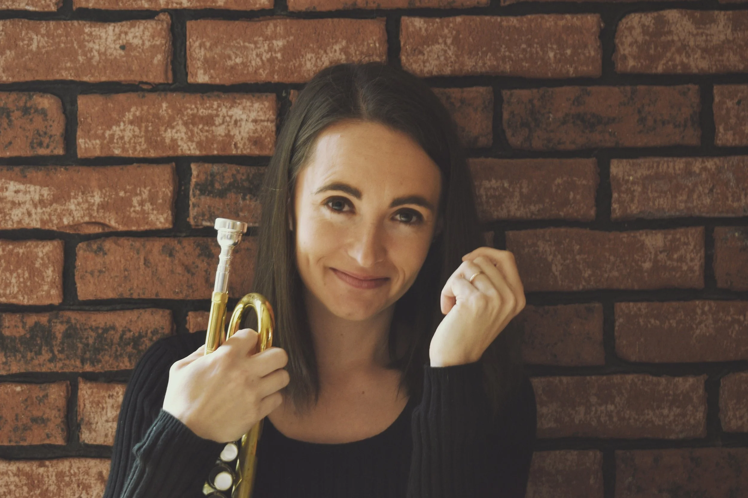 Woman holding a brass trumpet in front of a brick wall