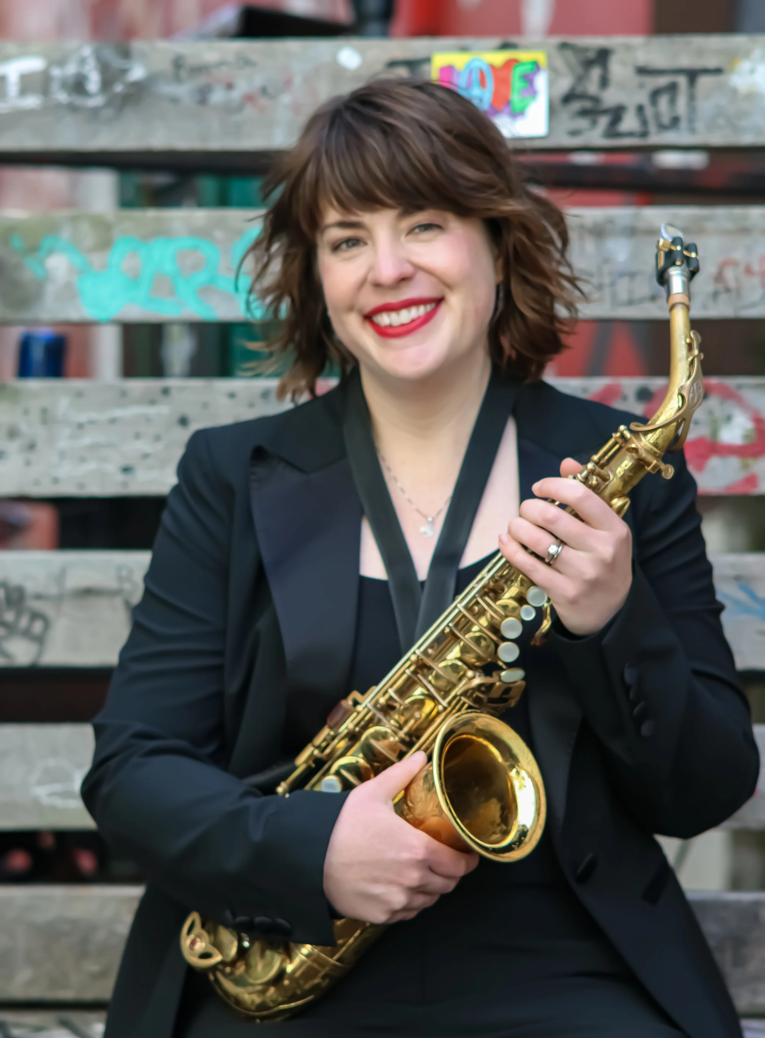 A woman with shoulder-length brown hair, wearing a black blazer and red lipstick, smiling while holding a gold-colored saxophone against a graffiti-covered metal staircase background.