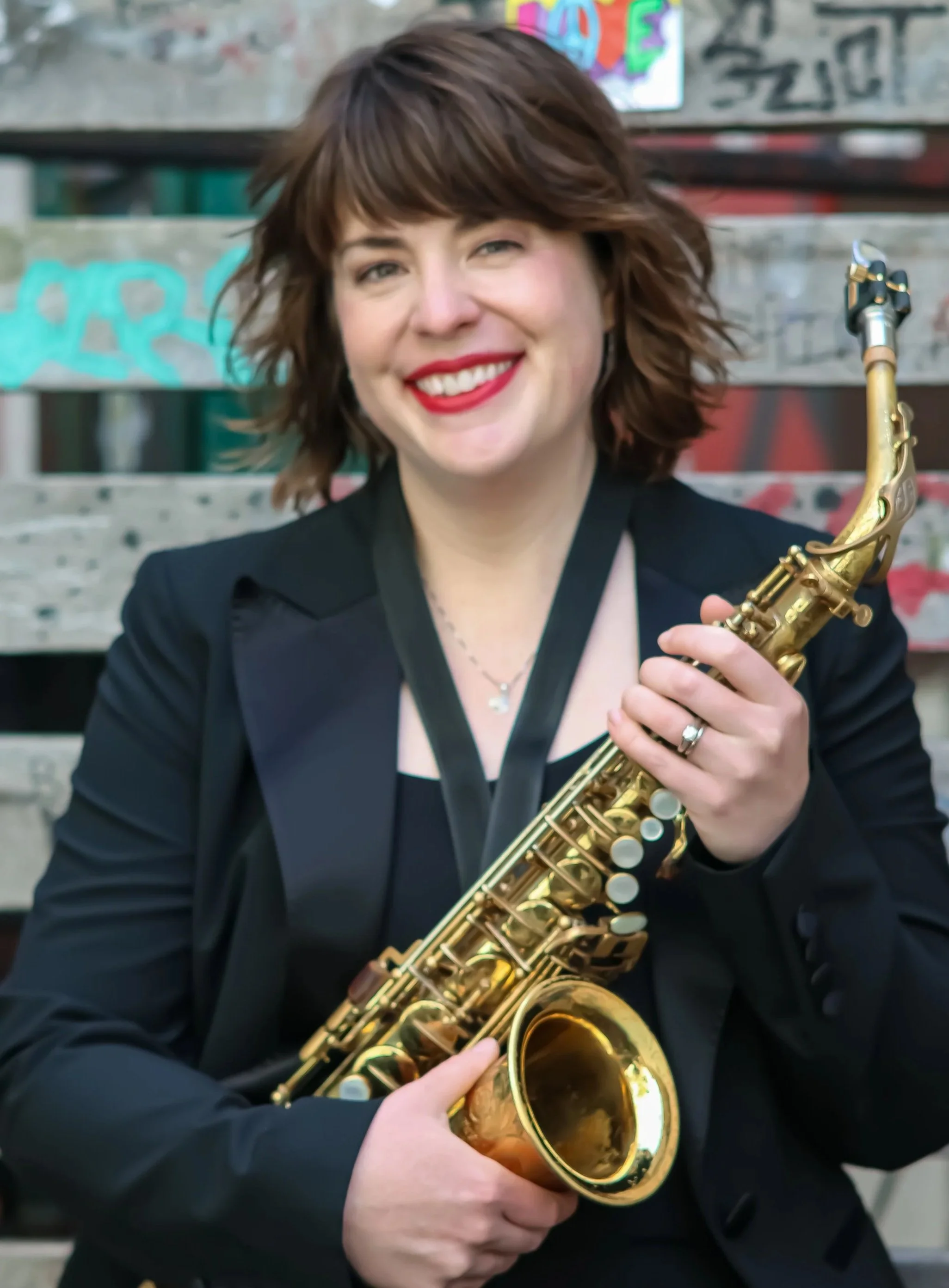 Smiling woman in a black blazer holding a gold saxophone in front of a graffiti-covered wall