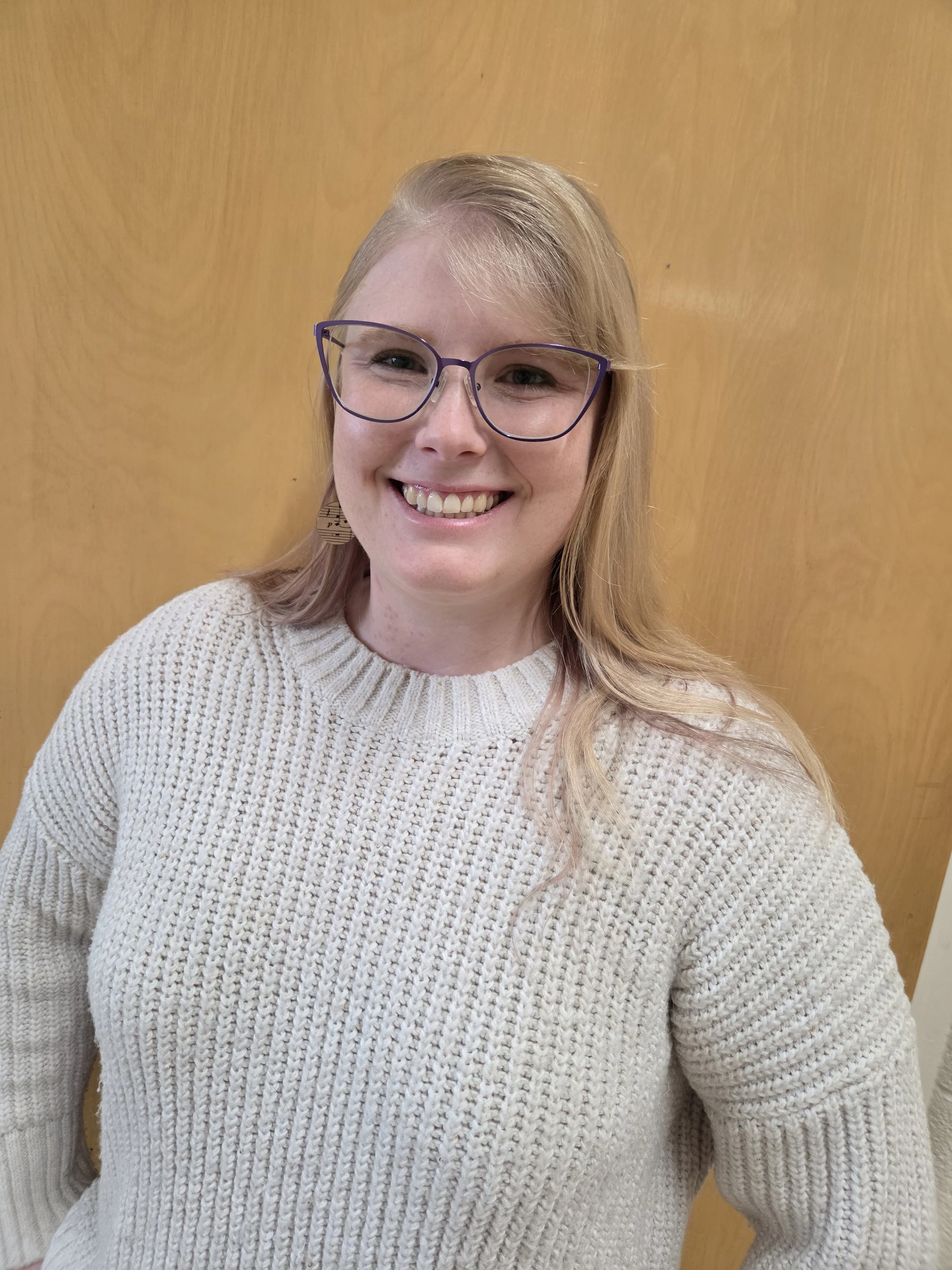 A smiling young woman with blonde hair, glasses, and a cream-colored knit sweater standing against a wooden background.
