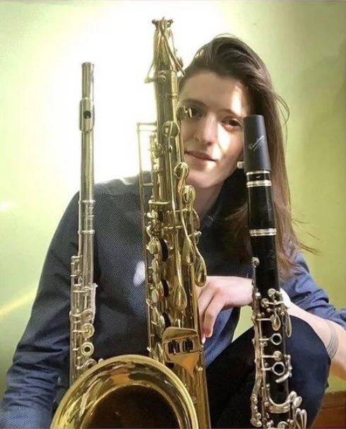 A woman with long brown hair holding a clarinet, sitting behind a saxophone and an oboe, against a light green wall.