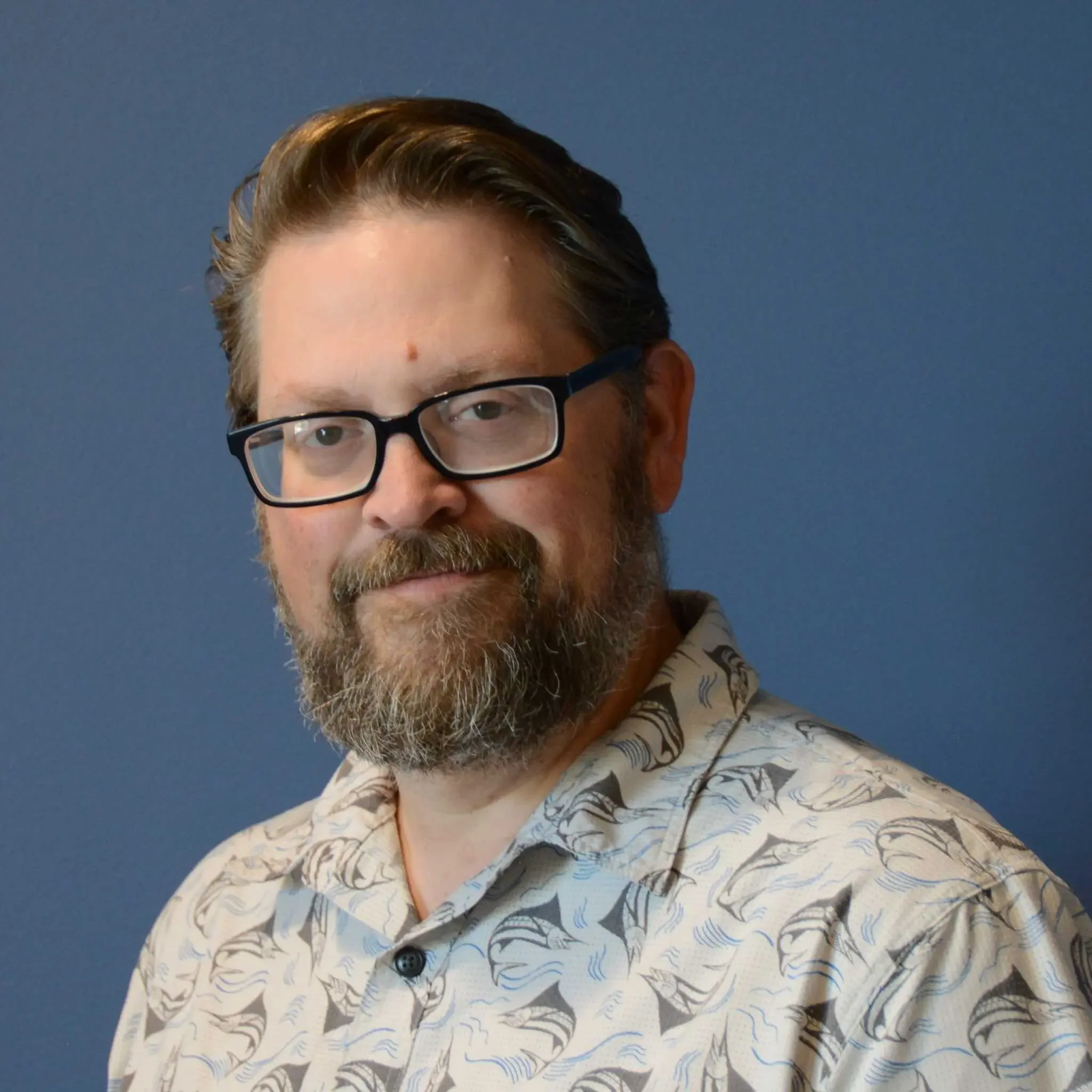 Portrait of a bearded man wearing black glasses and a patterned button-up shirt against a blue background