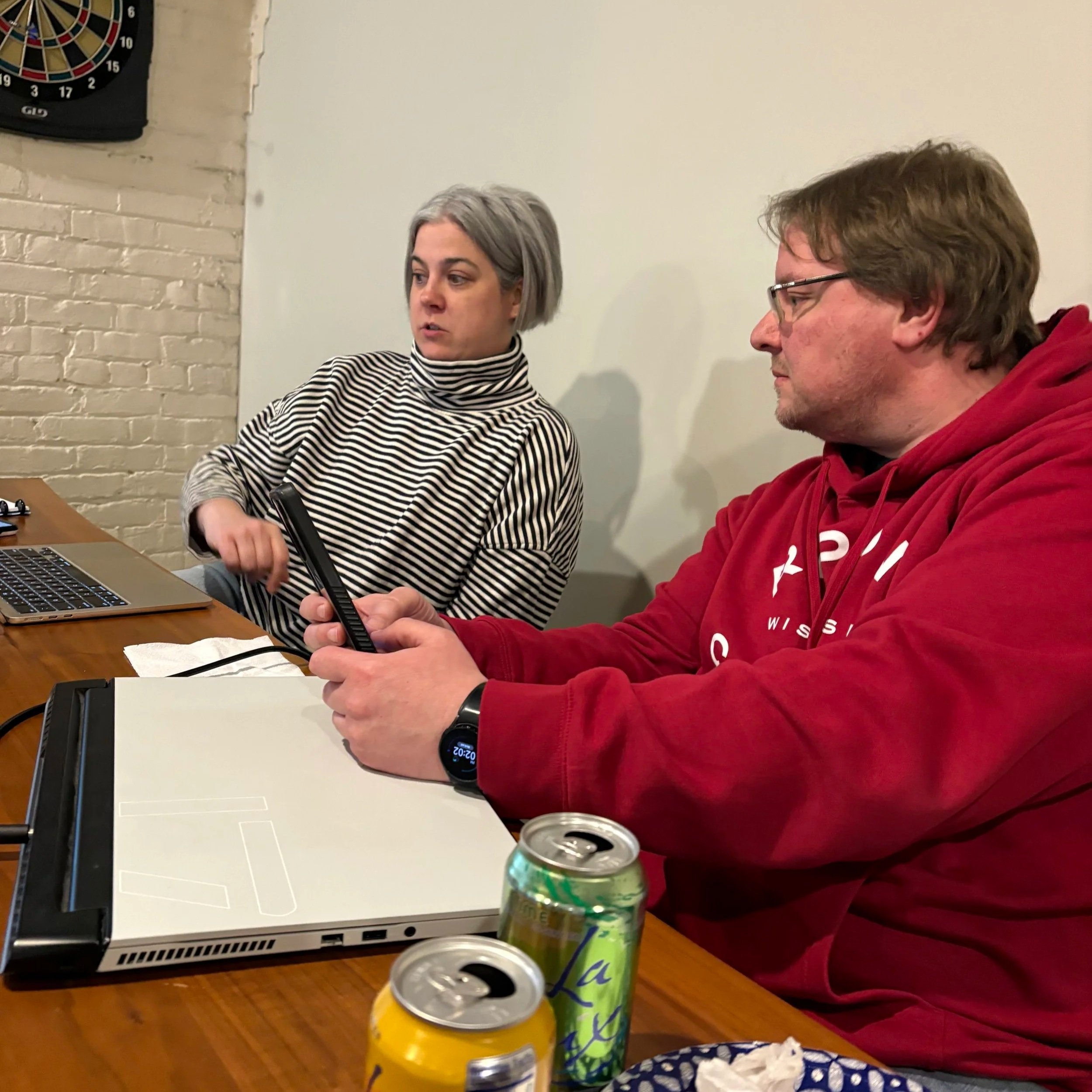 Two people sitting at a table, one woman with short gray hair wearing a black and white striped shirt, and one man with brown hair and glasses in a red hoodie, with a laptop, drink cans, a pizza box, and a dartboard on the wall in the background.